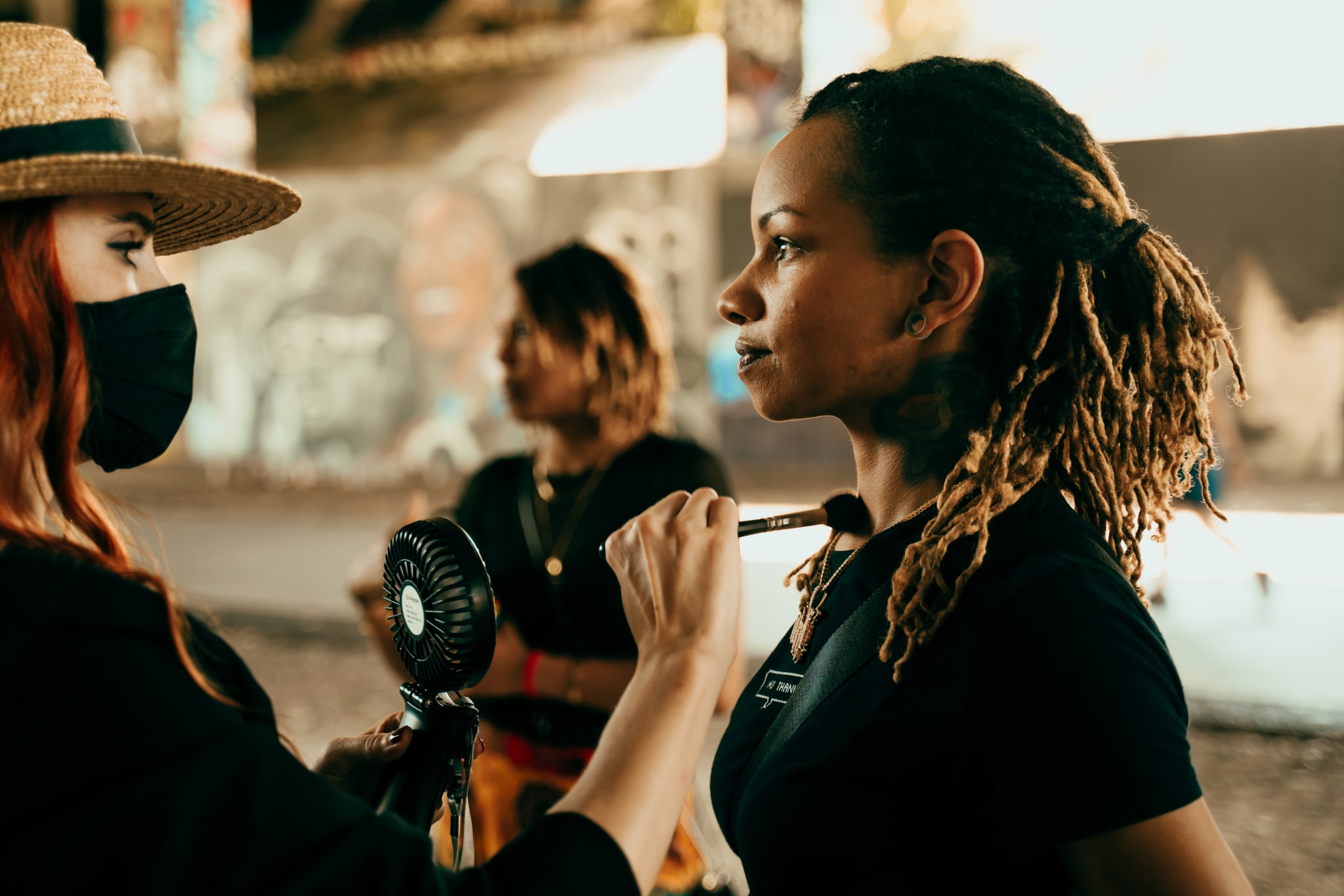 Three women, one with a straw hat and fan, apply makeup to another woman with dreadlocks in Atlanta, Georgia.
