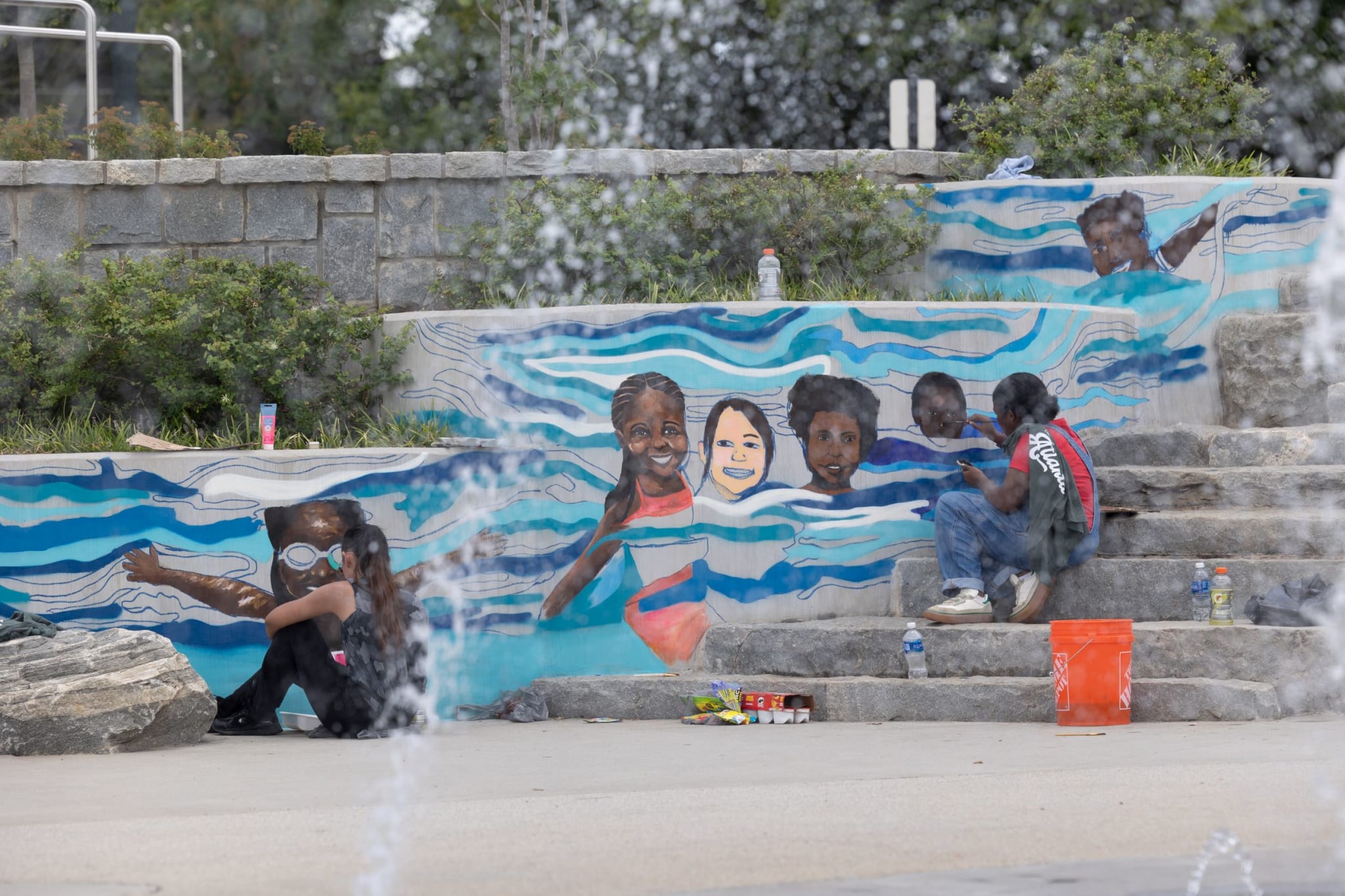 A mural of children swimming adorns a stone wall near a fountain, with people relaxing on the steps in Atlanta, Georgia.