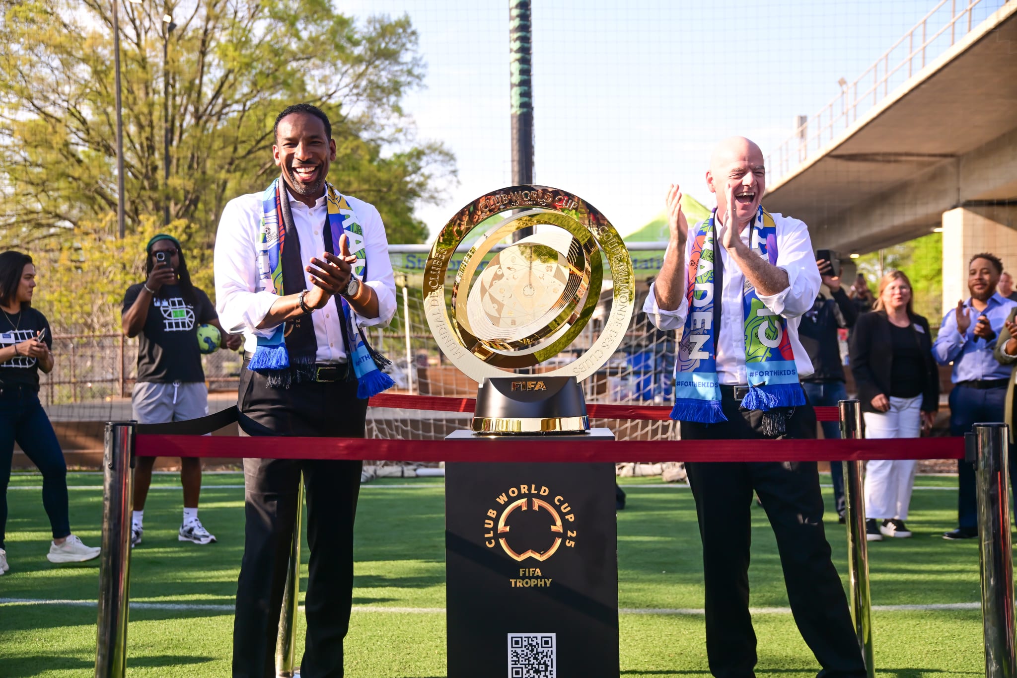 Two men in suits stand beside a large golden trophy on a soccer field, surrounded by cheering people in Atlanta, Georgia.