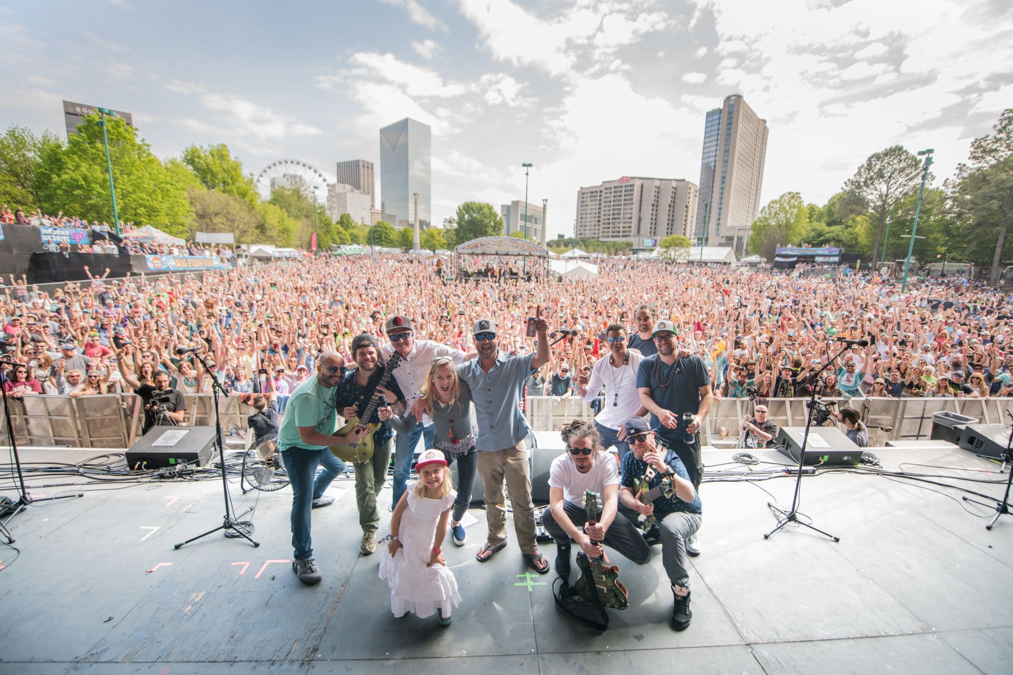 A large group of musicians and a child pose on stage in front of a massive, cheering crowd at an outdoor music festival in Atlanta, Georgia.