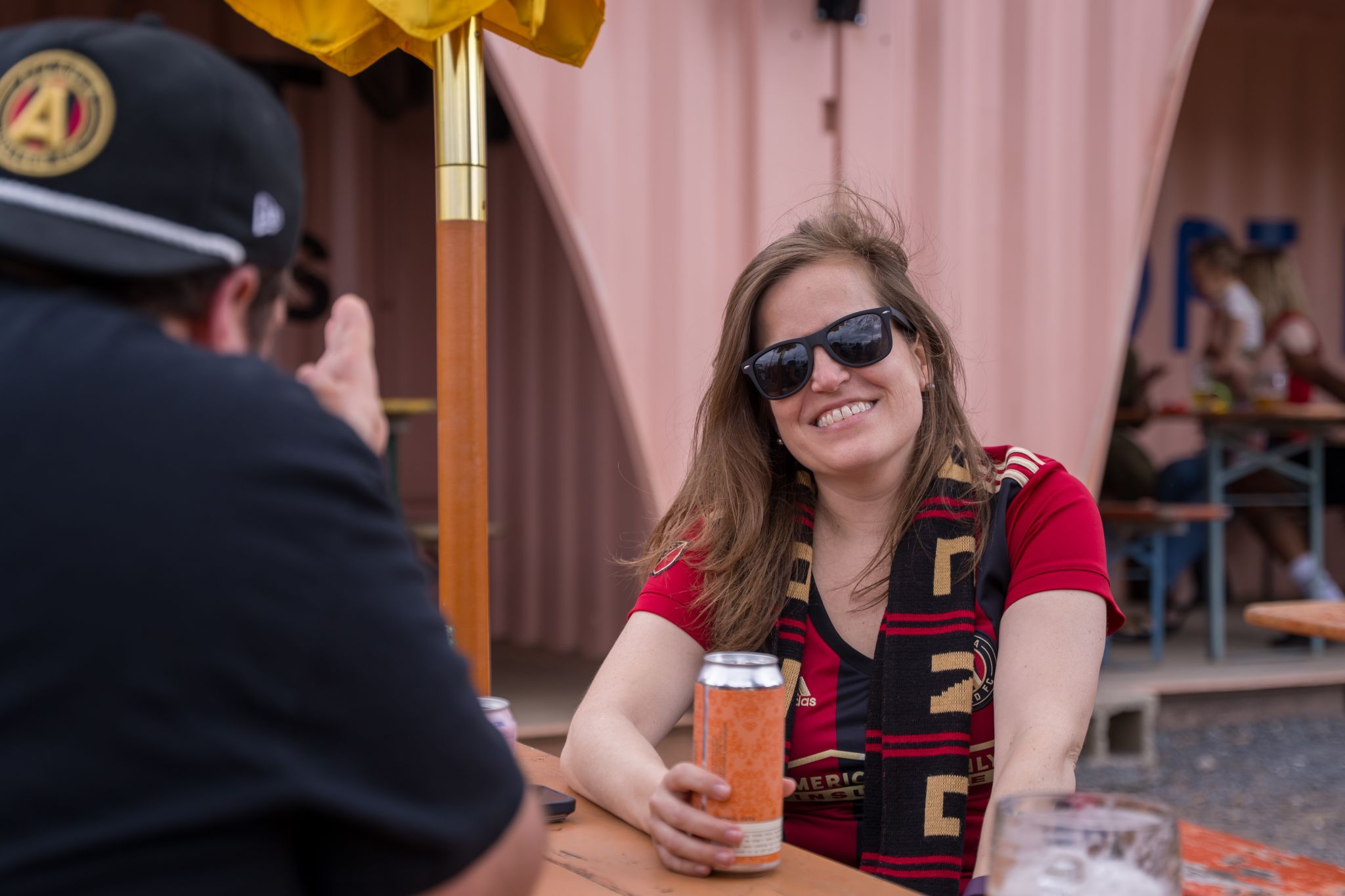A woman in a red soccer jersey and sunglasses enjoys a beer at an outdoor bar in Atlanta, Georgia.
