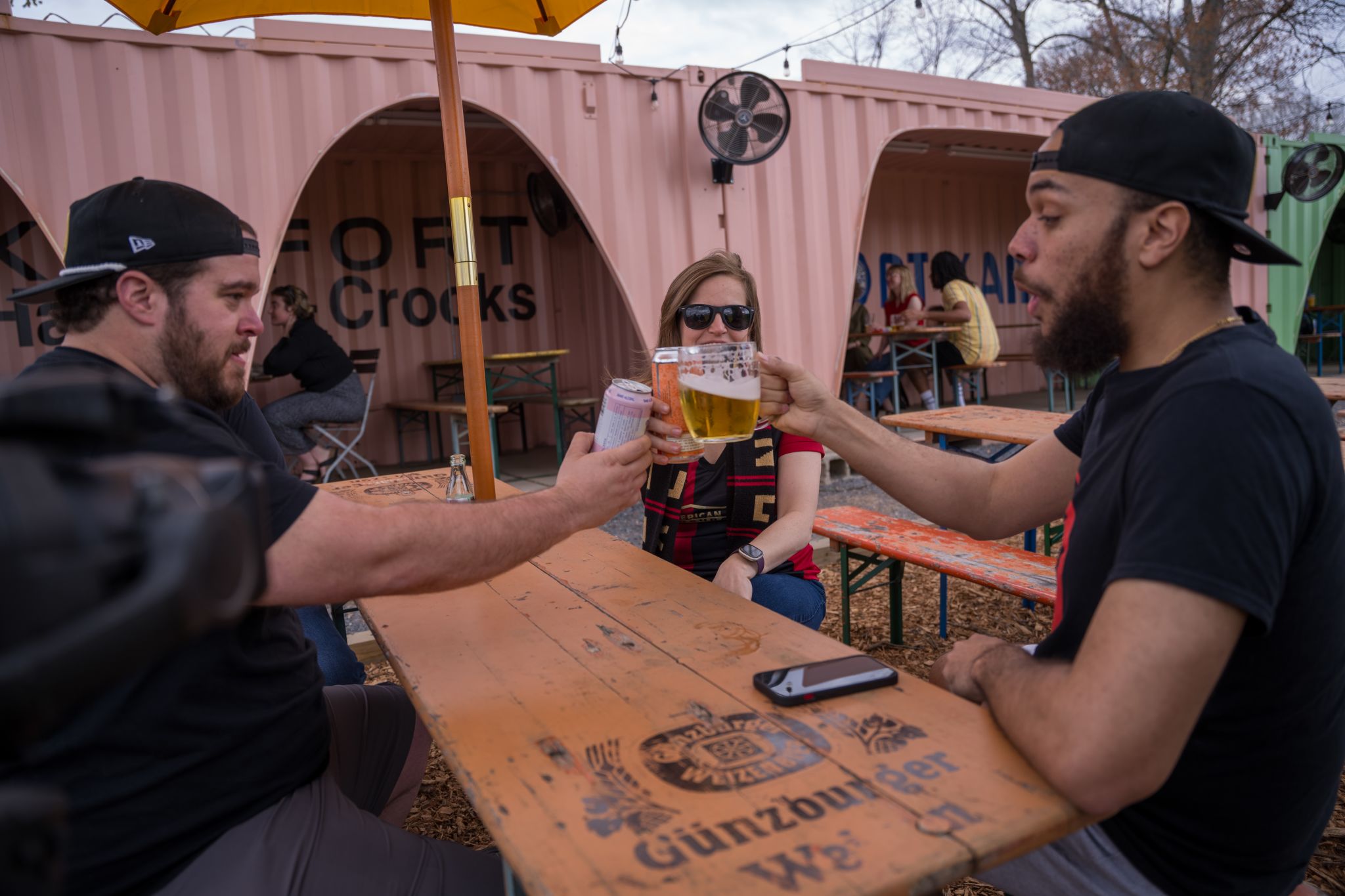 Three people clinking beer glasses at an outdoor picnic table under a pink container restaurant in Atlanta, Georgia.