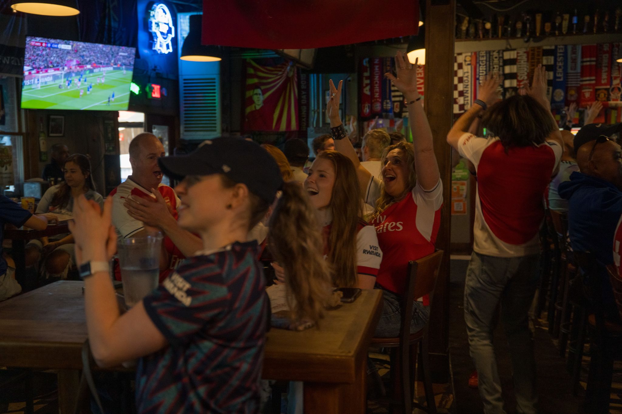Sports fans in a dimly lit bar cheer and clap while watching a soccer match on TV in Atlanta, Georgia.