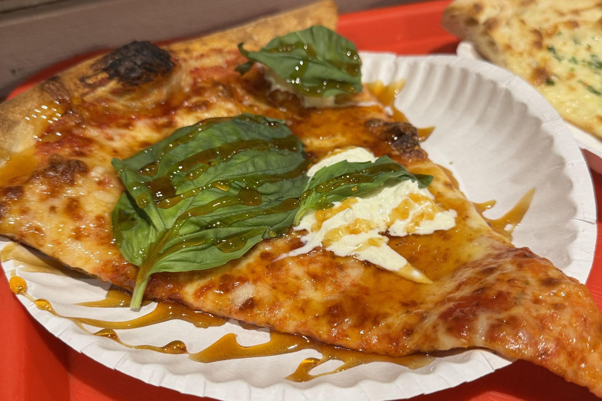 A slice of pizza topped with fresh basil leaves and dollops of creamy cheese sits on a white paper plate, drizzled with a golden syrup, all resting on a vibrant red tray in Atlanta, Georgia.