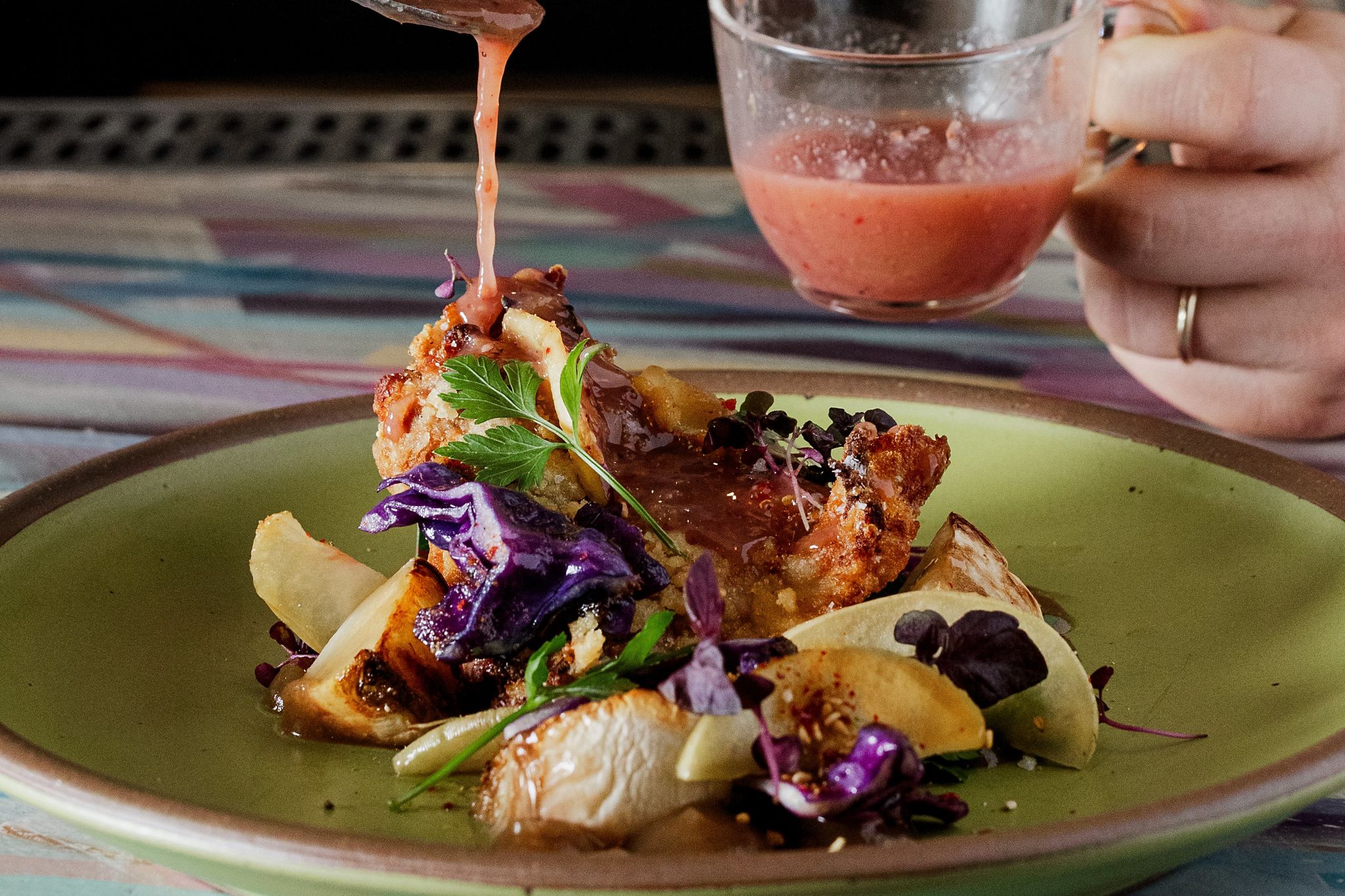 A hand pours a vibrant red sauce from a small glass cup onto a colorful dish of food, which includes crispy fried chicken, purple cabbage, and roasted vegetables, all served on a green plate in Atlanta, Georgia.