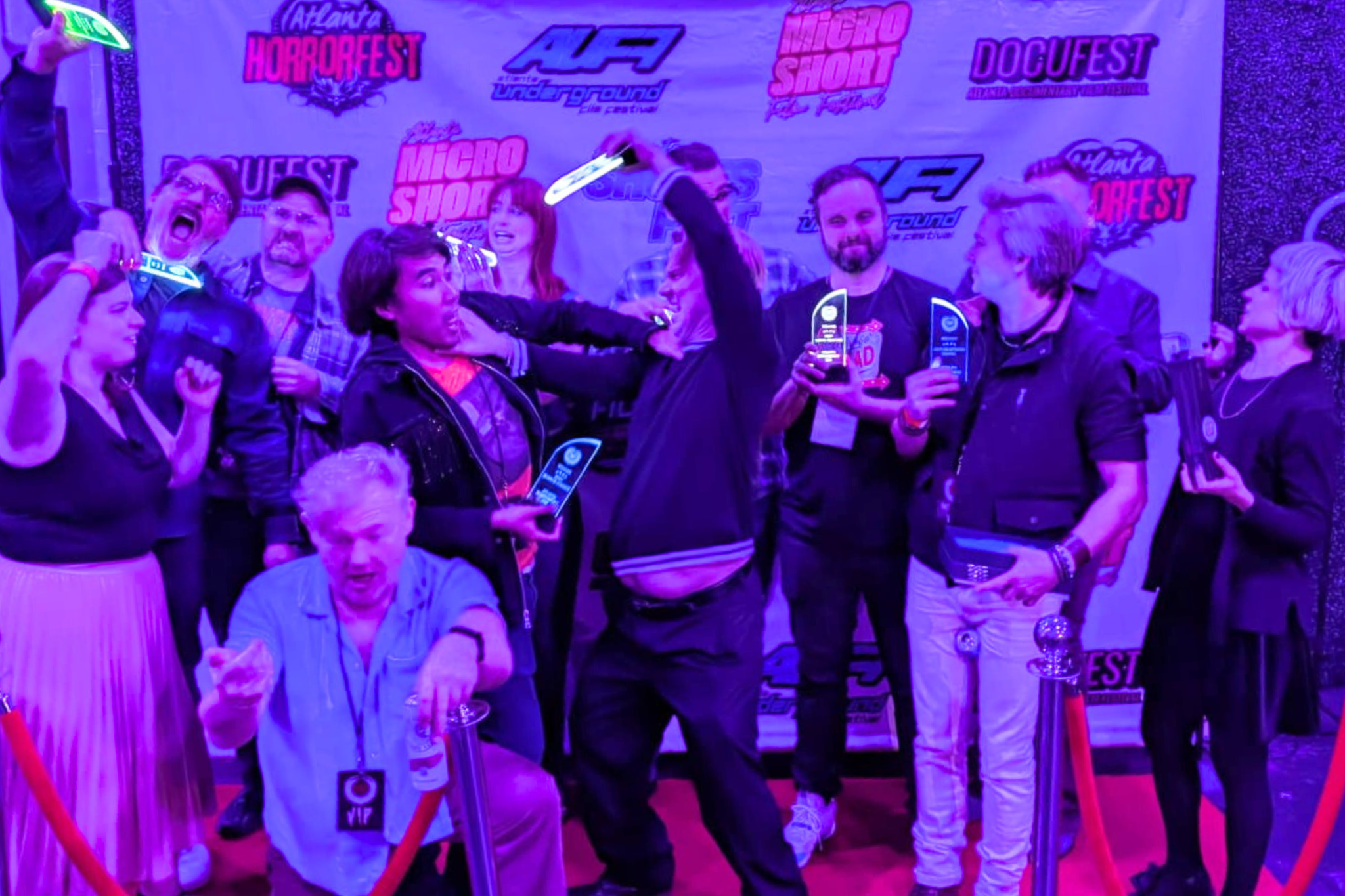 A group of enthusiastic people celebrate their achievements at an awards ceremony, holding up their trophies and glowing wands against a backdrop of vibrant festival banners in Atlanta, Georgia.