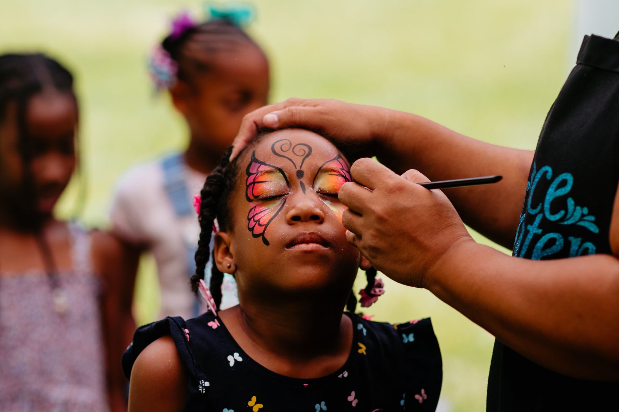 A young girl with braids sits still while an adult paints intricate butterfly face paint on her eyes in Atlanta, Georgia.