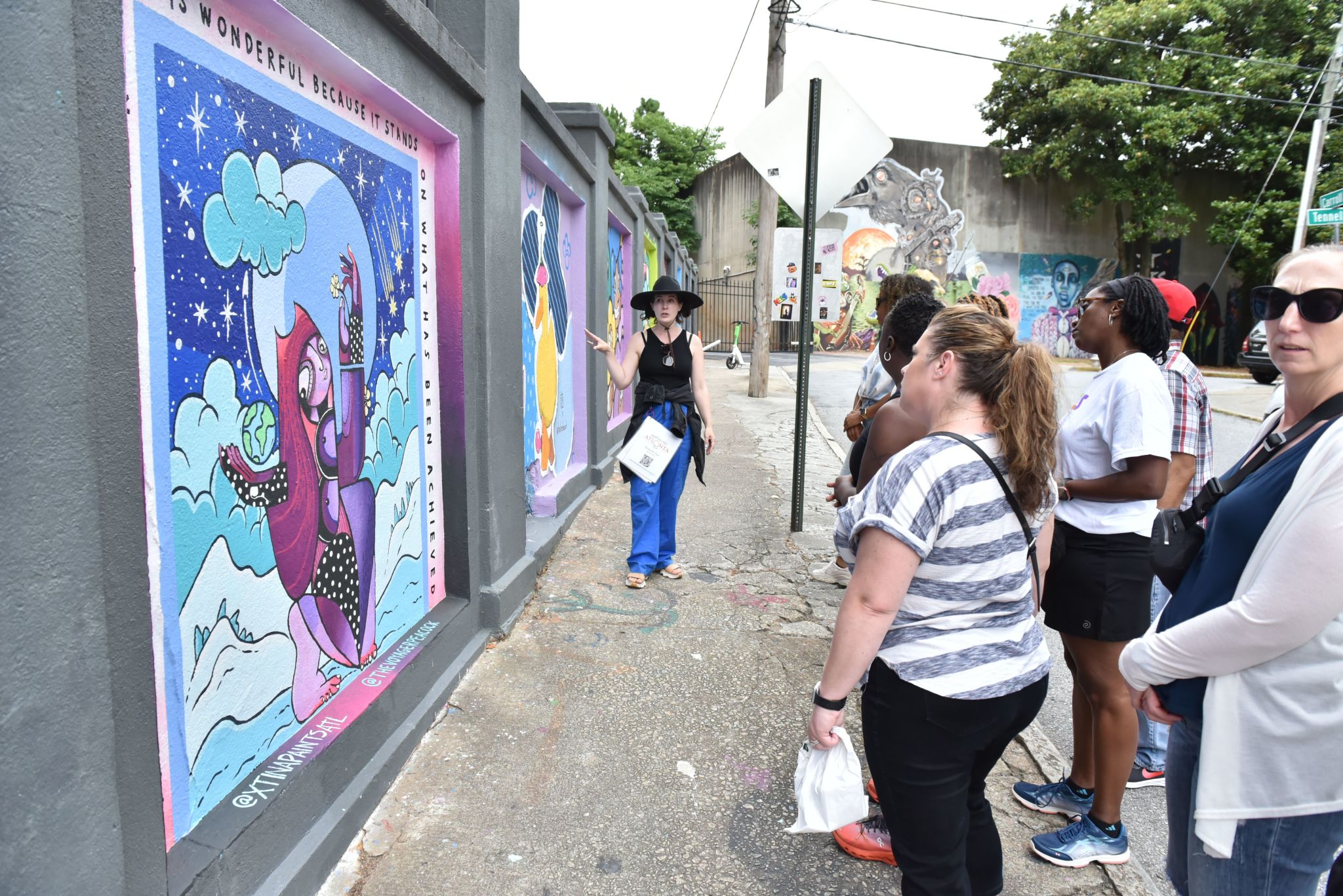 A group of people are gathered on a sidewalk, admiring a vibrant mural on a wall that depicts a whimsical scene with a woman in a purple dress and a starry night sky in Atlanta, Georgia.