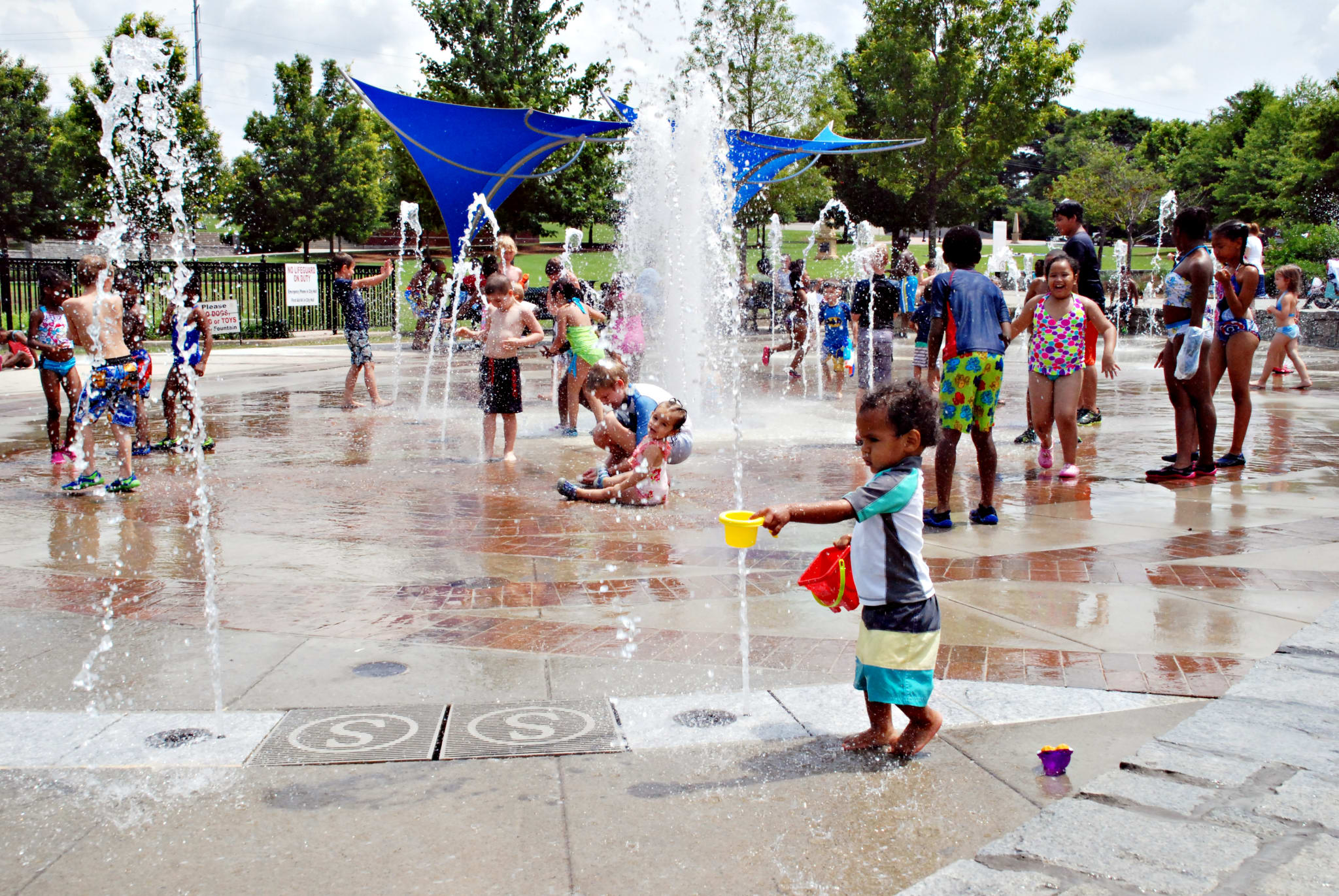Children play in a splash pad with multiple water fountains and blue shade structures in Atlanta, Georgia.