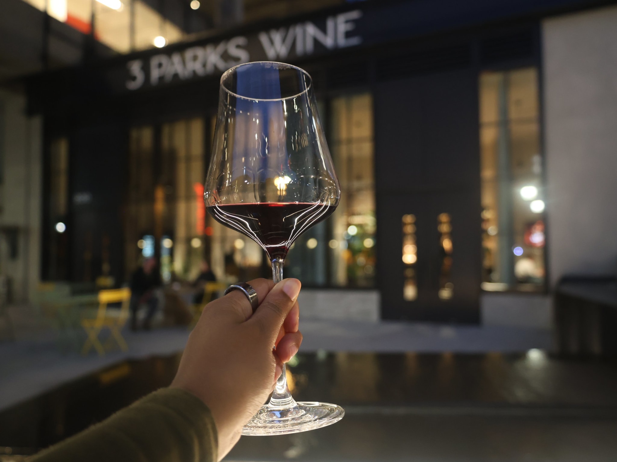 A hand holds a glass of red wine in front of a dimly lit restaurant named '3 Parks Wine' in Atlanta, Georgia.