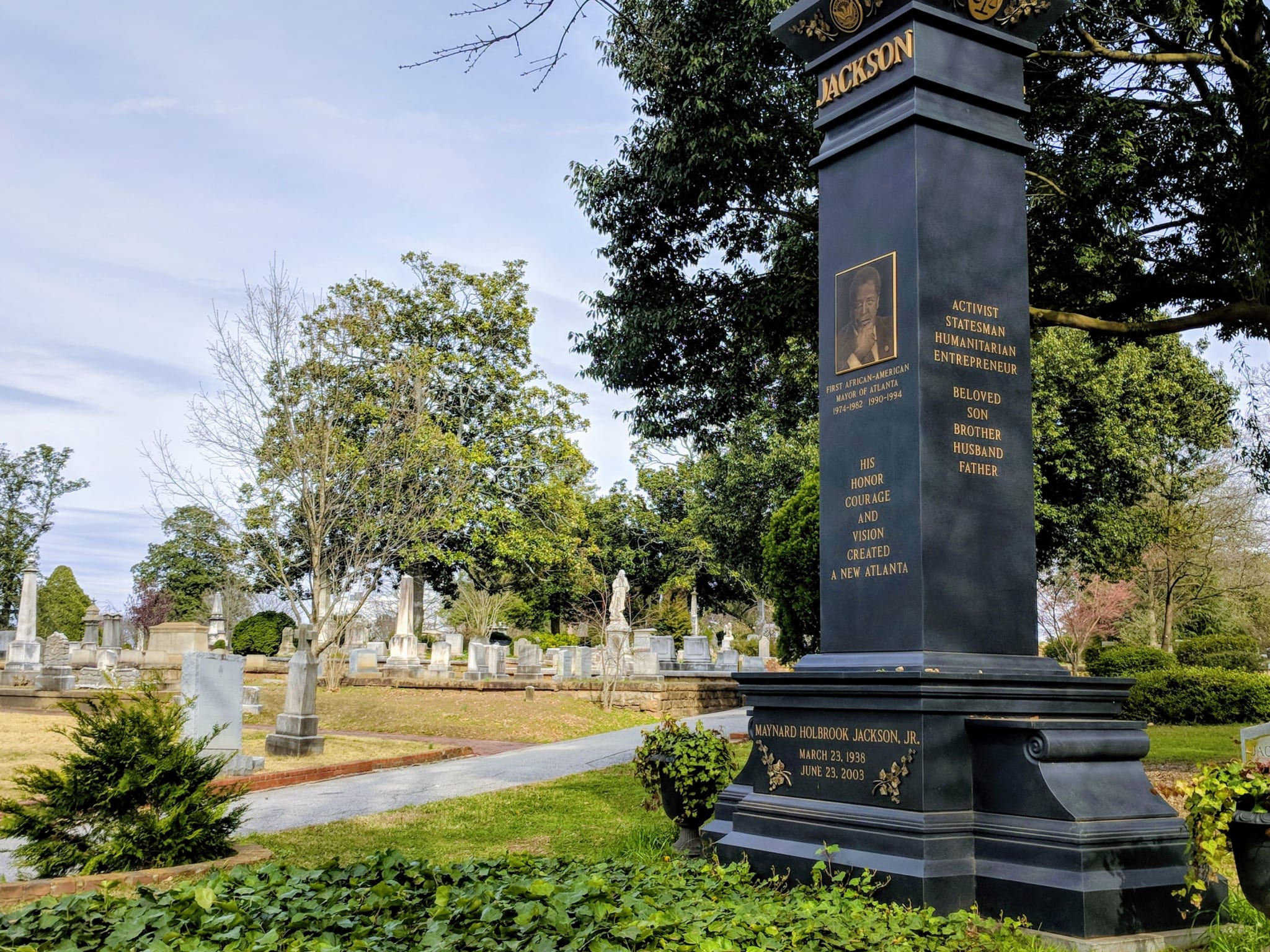 A tall, dark monument with a portrait and inscriptions stands in a serene cemetery with trees and gravestones in Atlanta, Georgia.
