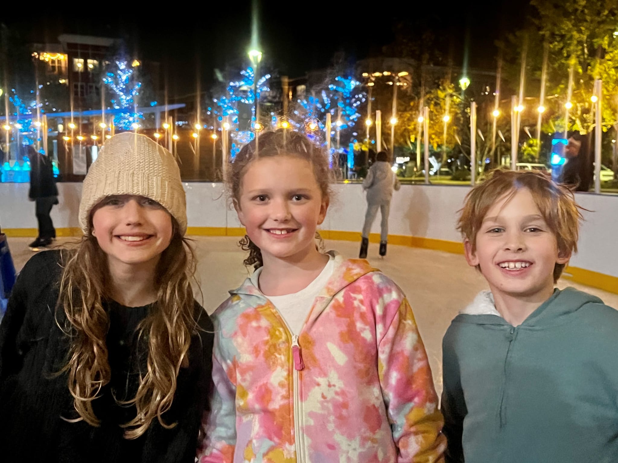 Three smiling children wearing winter clothing stand together on an ice rink at night in Atlanta, Georgia.