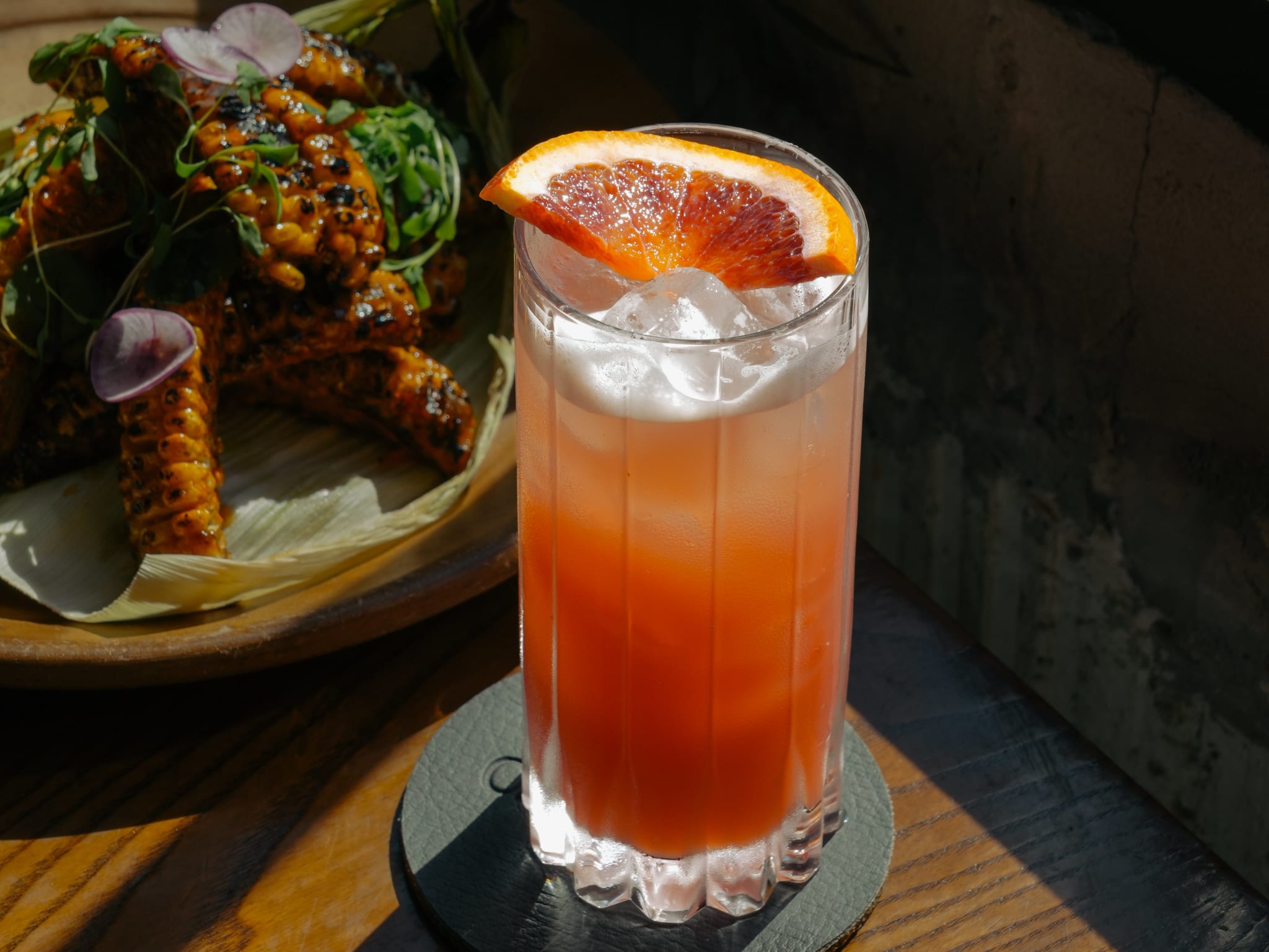 A refreshing pink cocktail garnished with an orange slice sits on a wooden table next to a plate of grilled corn on the cob in Atlanta, Georgia.