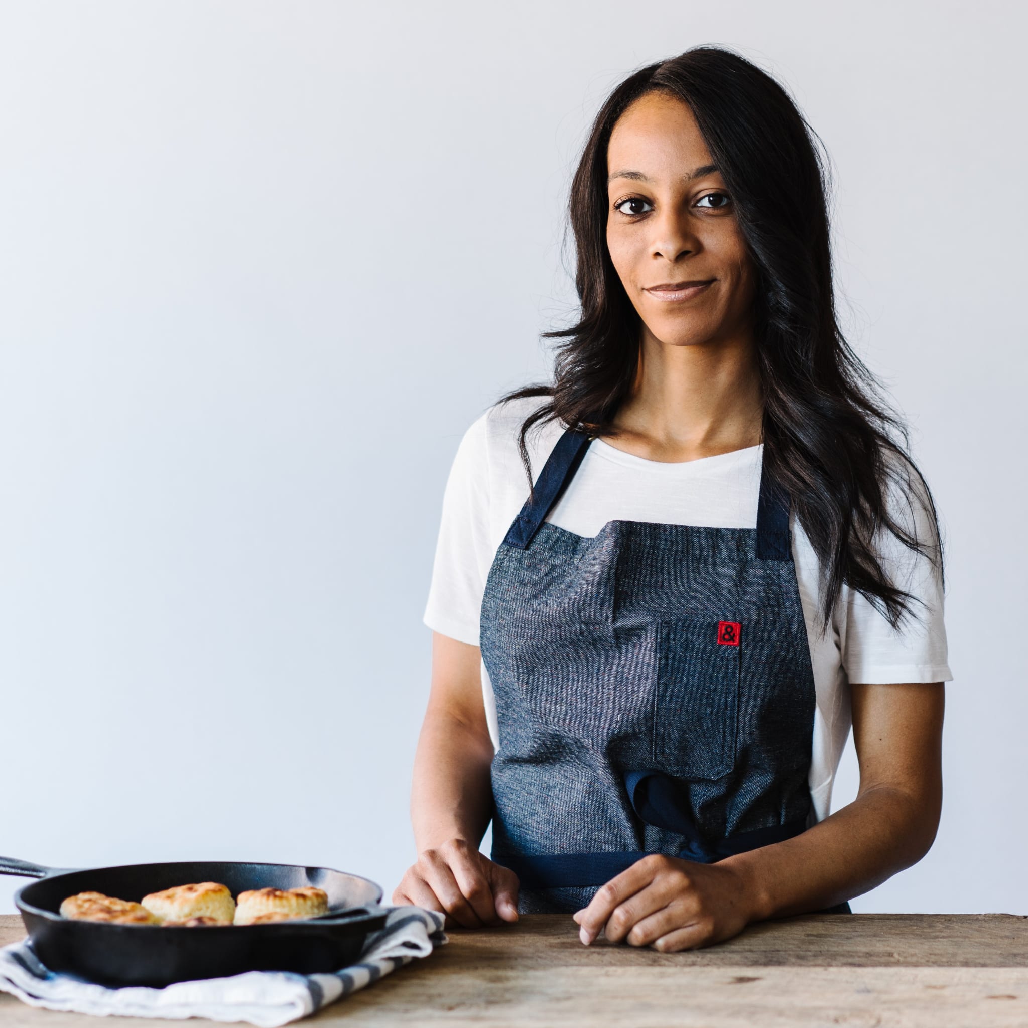 A woman wearing a denim apron stands behind a wooden table with a cast iron skillet containing golden-brown biscuits in Atlanta, Georgia.