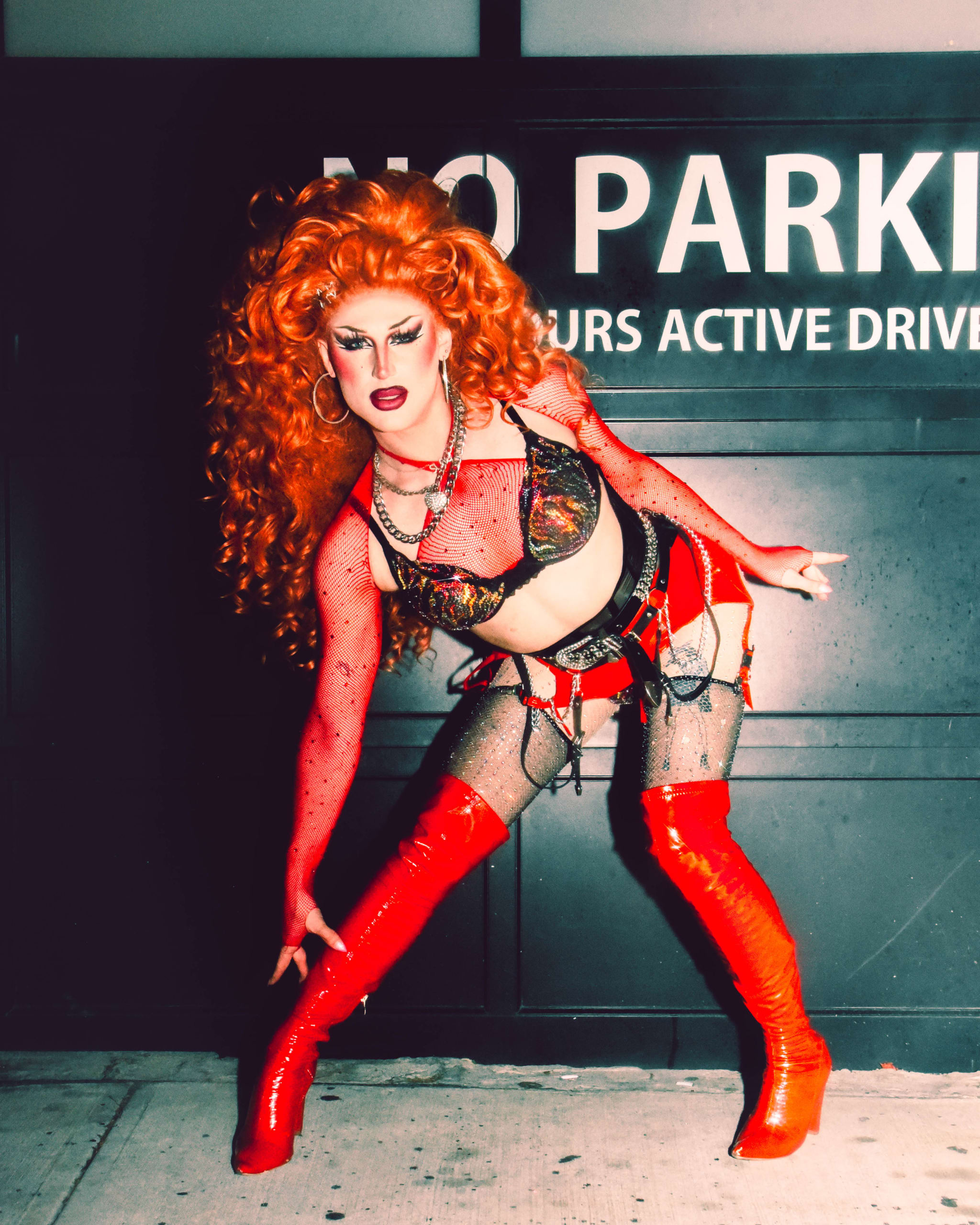 A person with vibrant red hair and makeup poses in a revealing red outfit against a parking garage backdrop in Atlanta, Georgia.