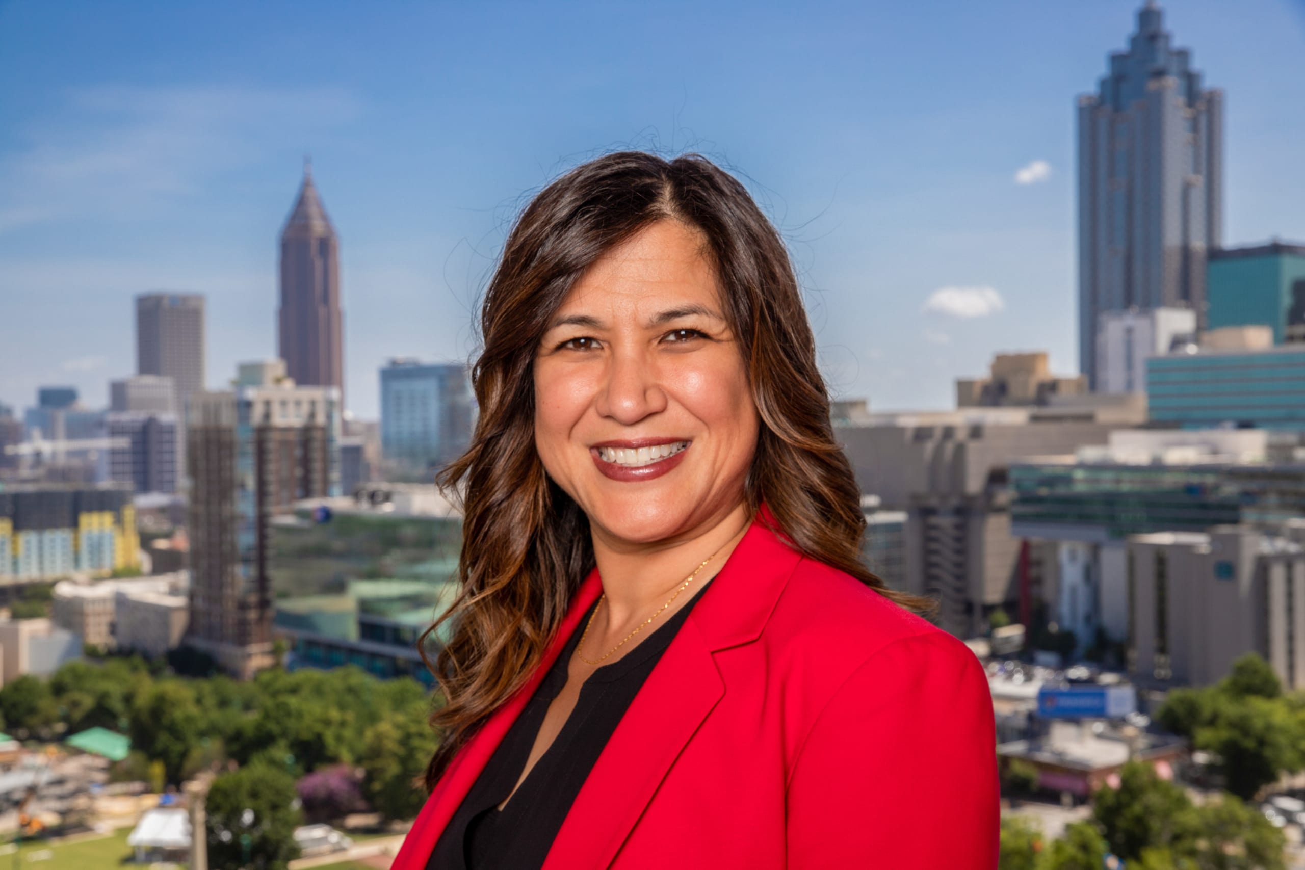 ACVB Promotes Charlene Lopez to Vice President 1 A smiling woman in a red blazer stands against a backdrop of a city skyline with tall buildings in Atlanta, Georgia.
