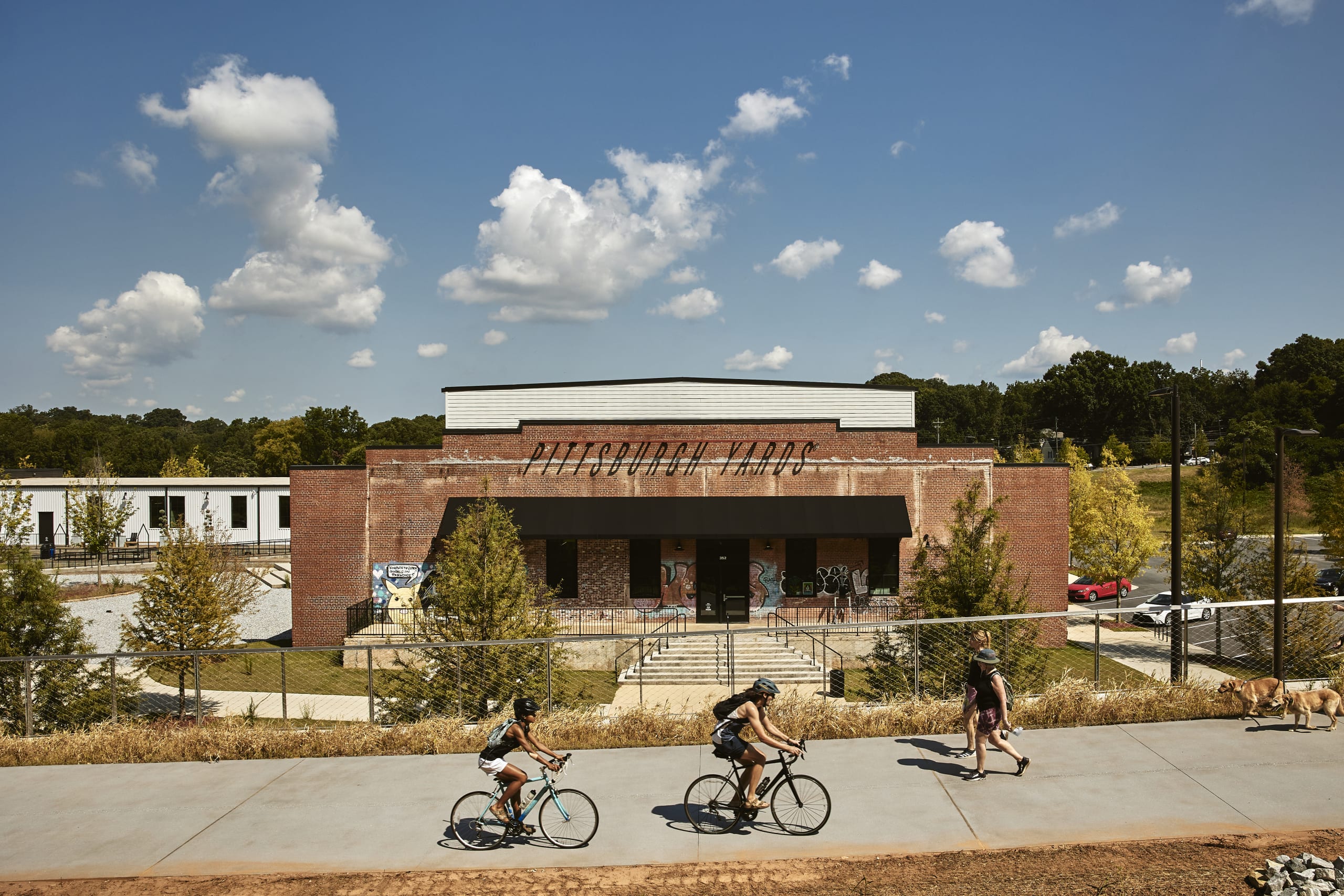Three cyclists and two pedestrians with dogs ride past a brick building with graffiti in Atlanta, Georgia.