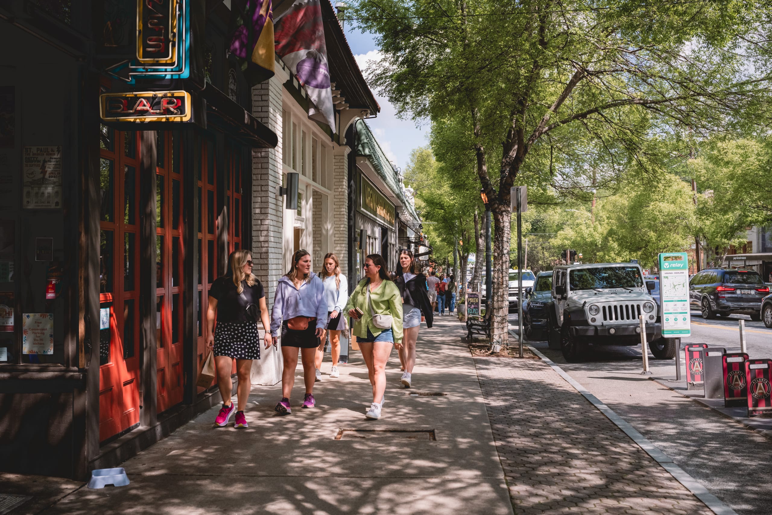 A group of women walk down a tree-lined sidewalk past shops and parked cars in Atlanta, Georgia.