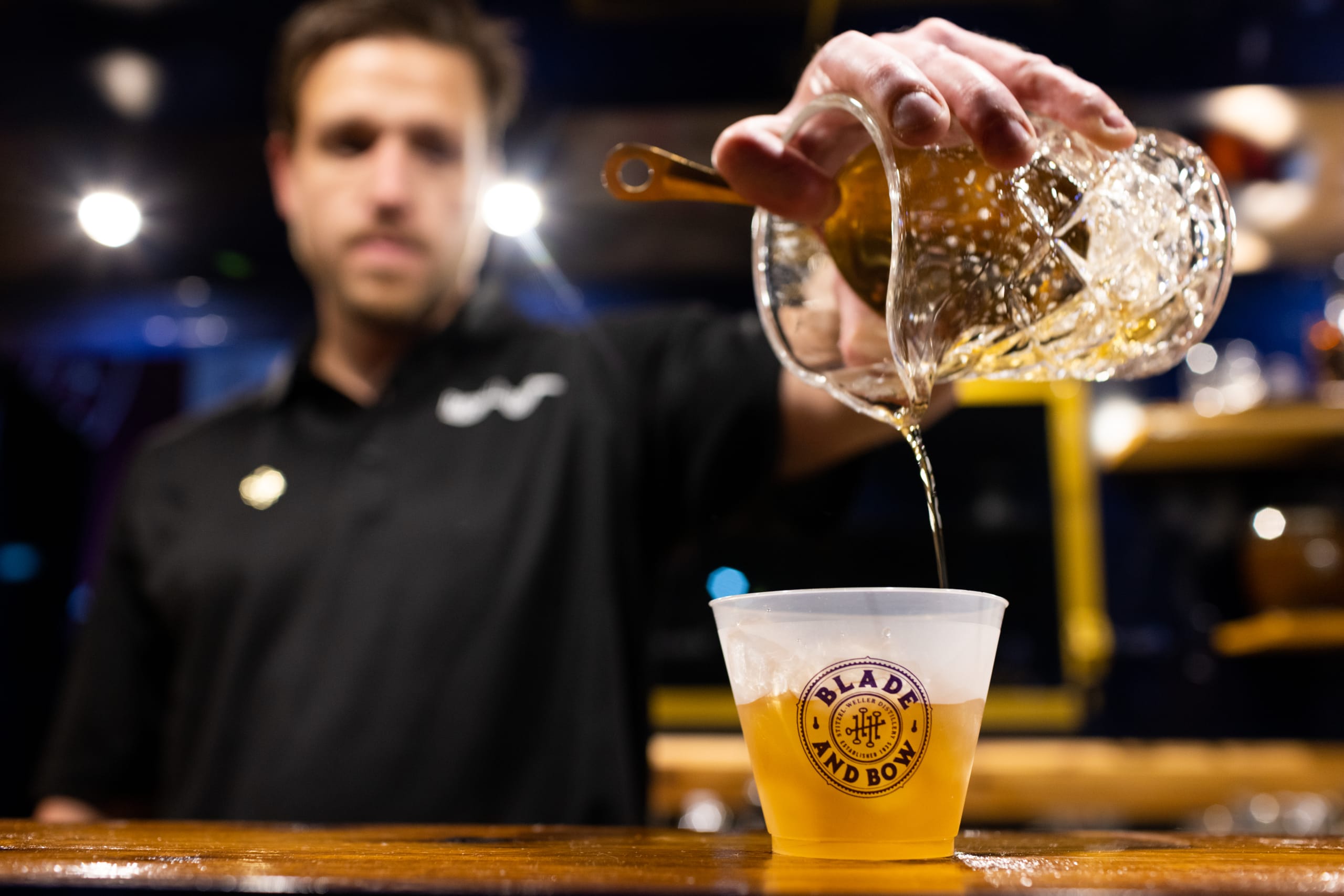 A bartender pours a golden liquid from a shaker into a plastic cup with a logo in Atlanta, Georgia.