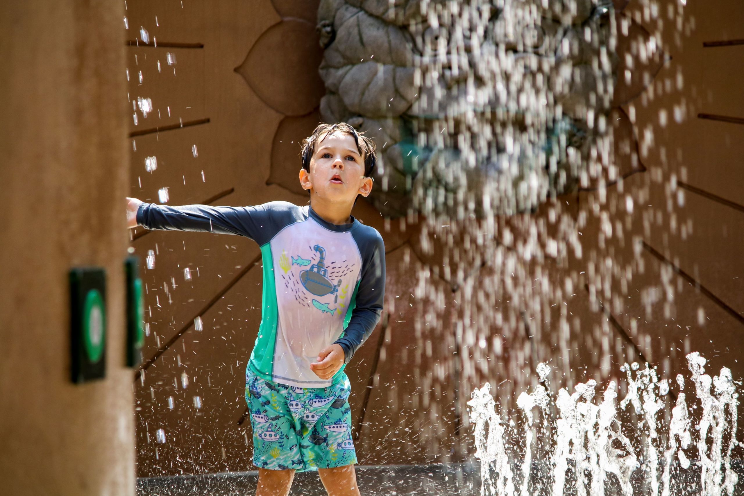 A young boy in colorful swimwear joyfully plays under a water fountain spray in Atlanta, Georgia.