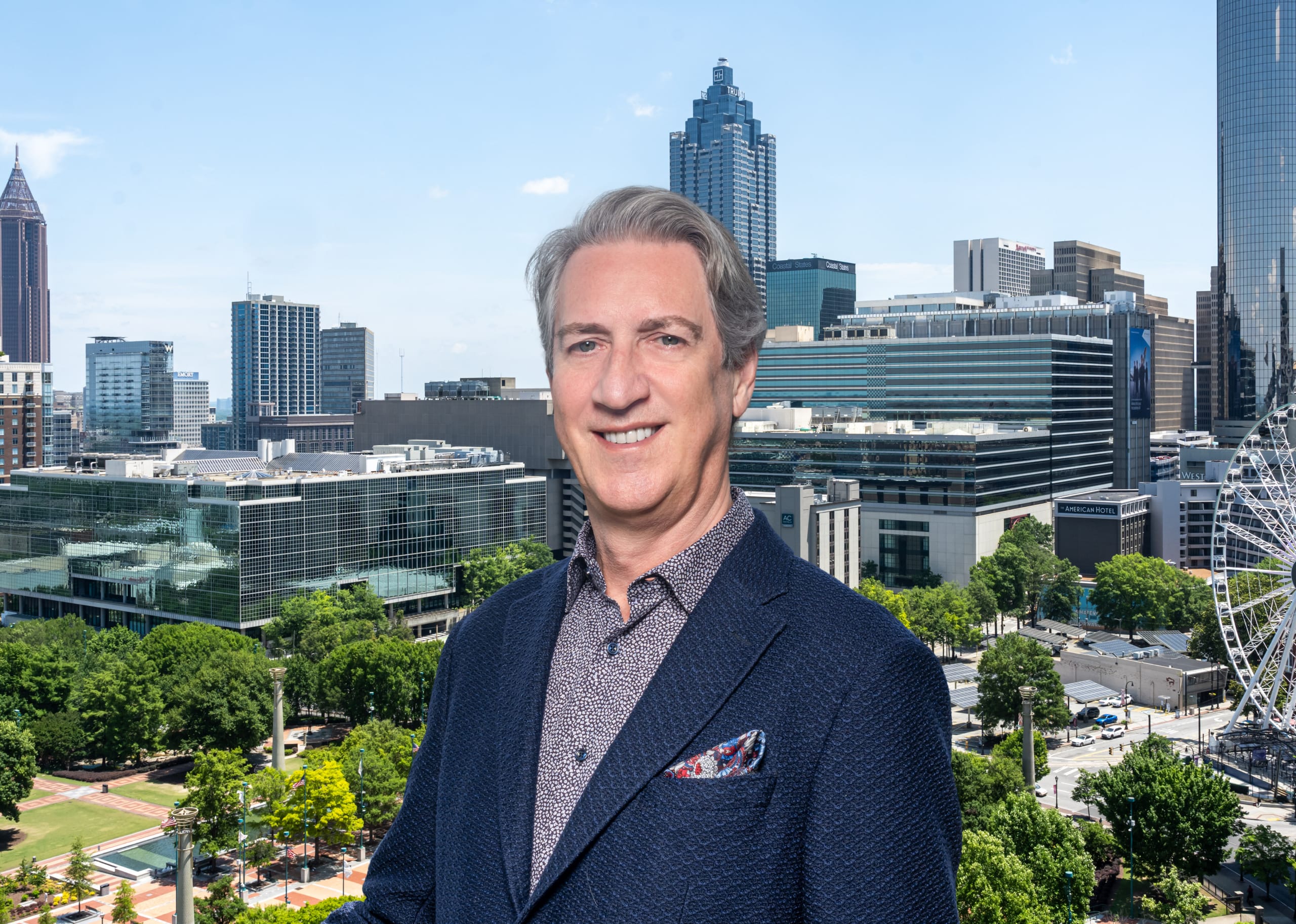 A man in a blue suit stands in front of a city skyline with skyscrapers and a Ferris wheel in Atlanta, Georgia.