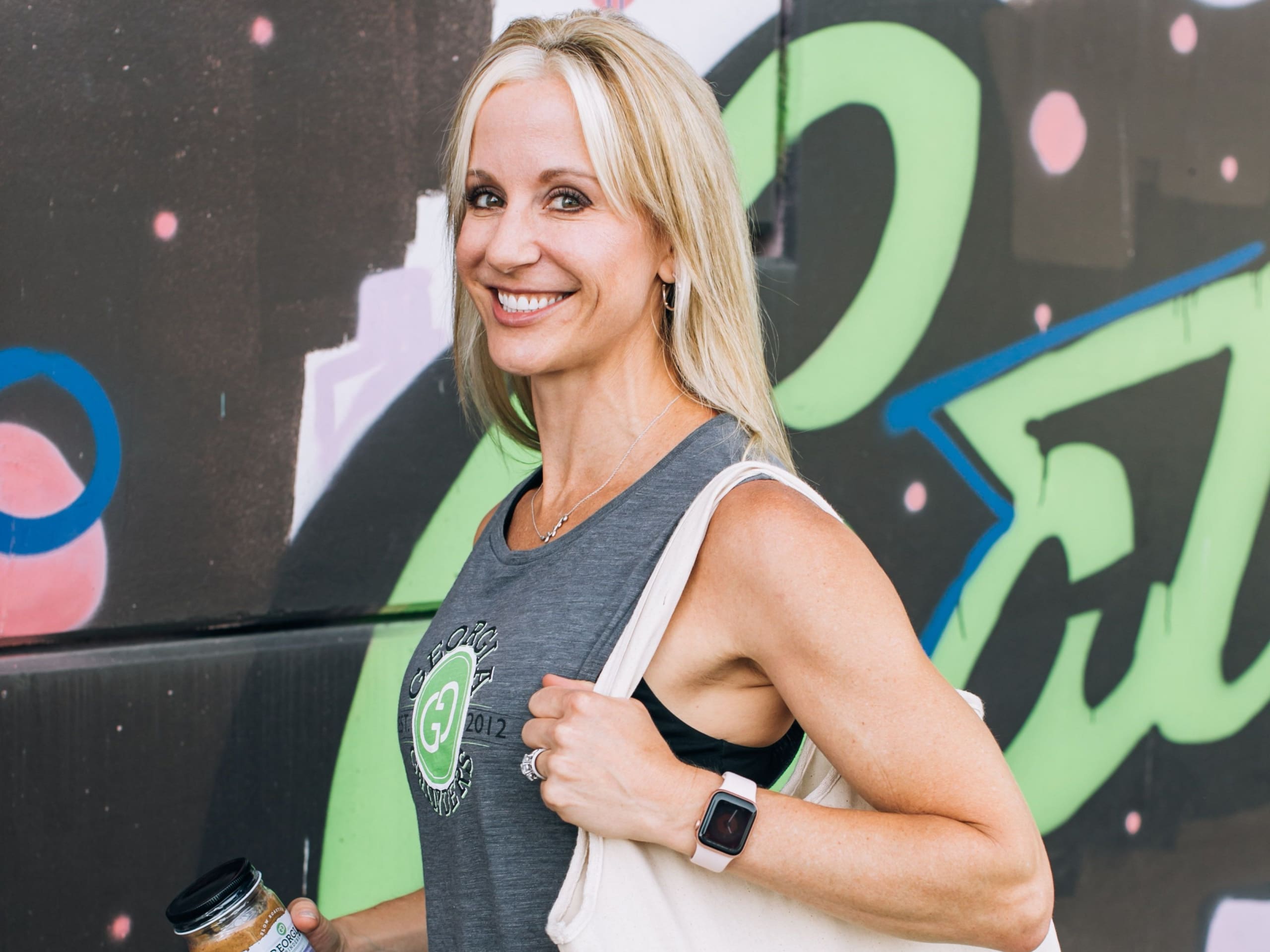 A smiling woman with blonde hair wearing a tank top and carrying a tote bag stands against a colorful graffiti wall in Atlanta, Georgia.