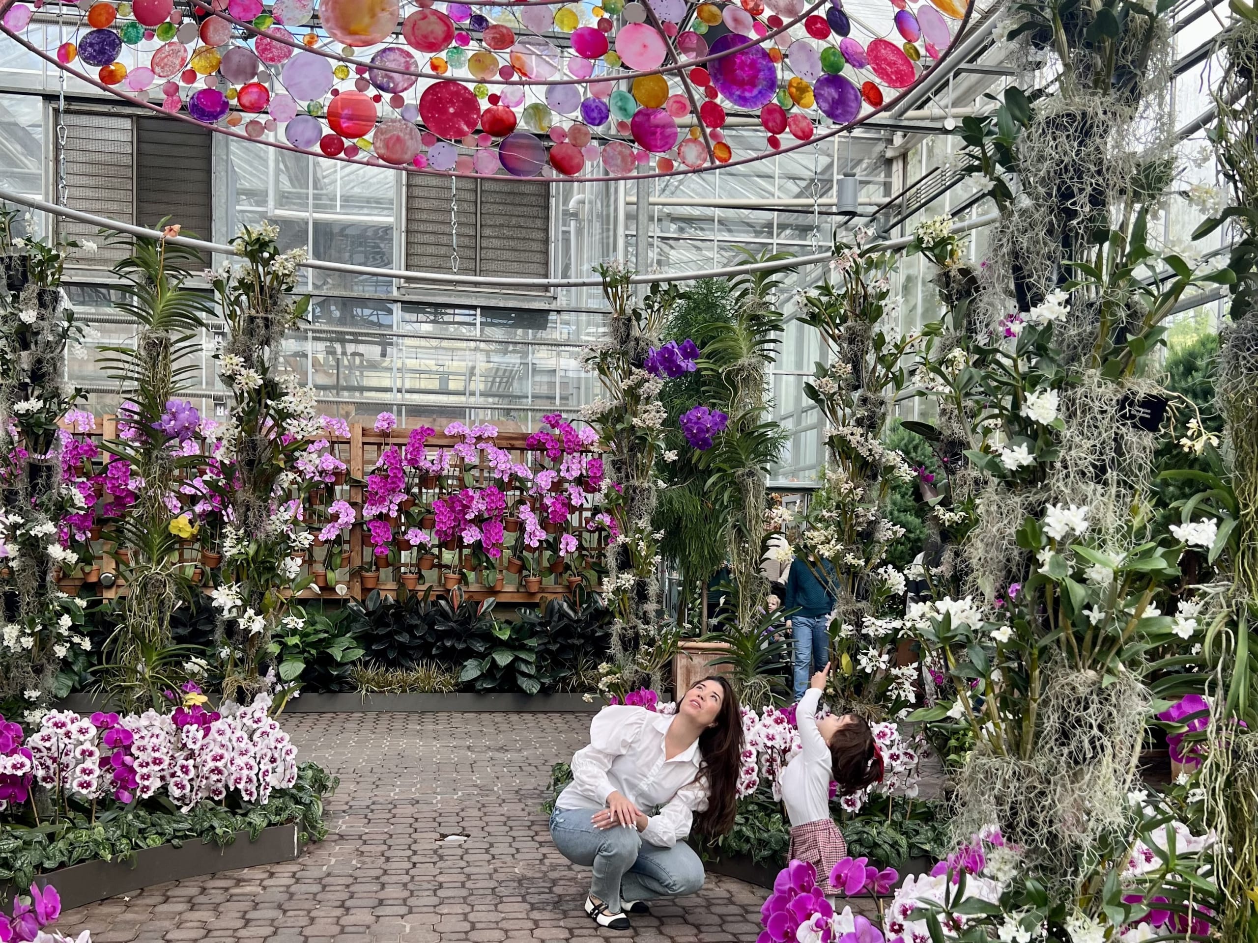 A woman and child kneel amidst a vibrant indoor garden filled with colorful flowers and lush greenery in Atlanta, Georgia.