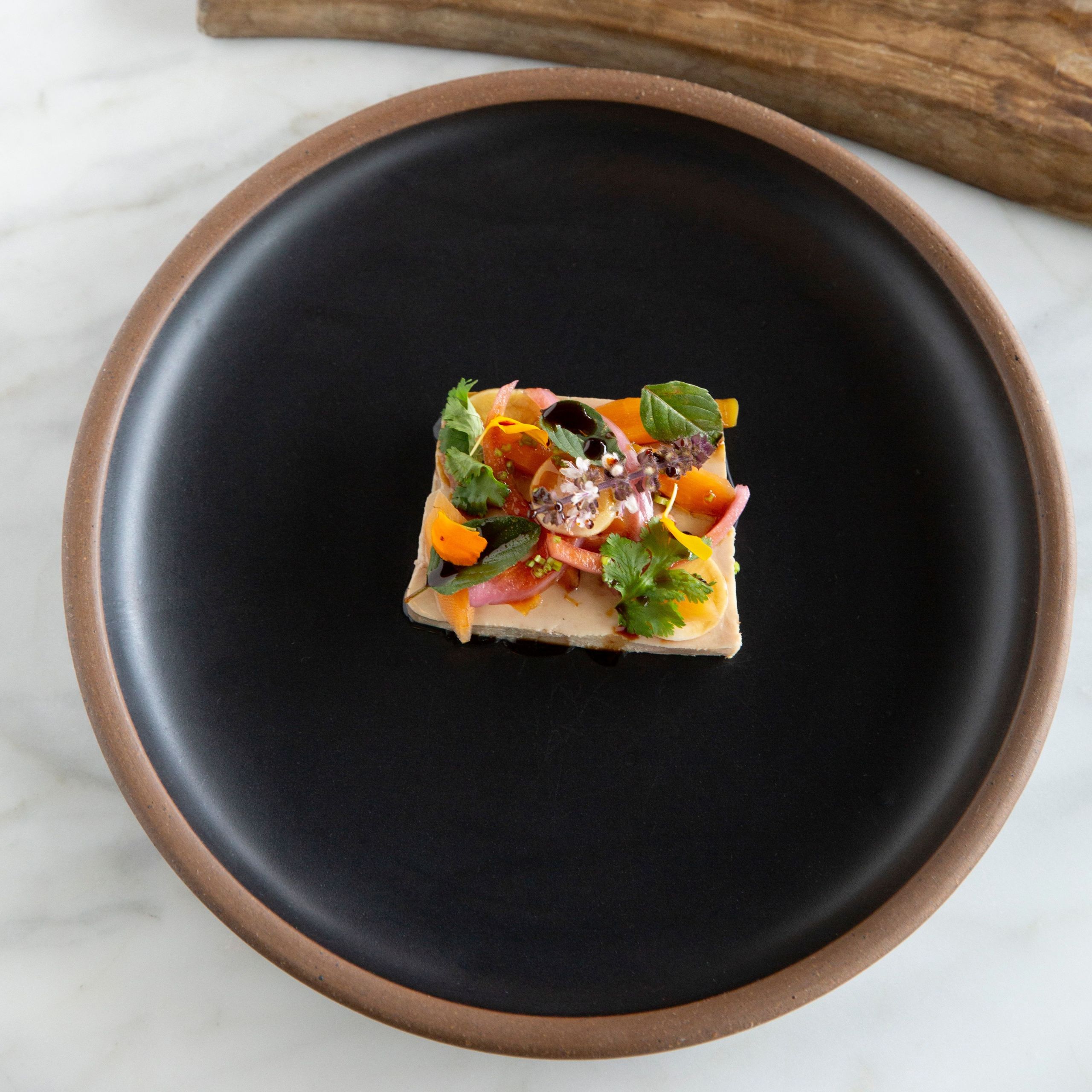 A square plate holds a colorful vegetable and herb garnish atop a creamy base in Atlanta, Georgia.