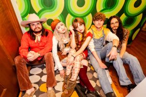 Ultimate Guide to Atlanta's Indie Music Scene 8 A group of five friends, dressed in colorful 70s-inspired outfits, sit together on a patterned rug against a vibrant green and yellow background in Atlanta, Georgia.
