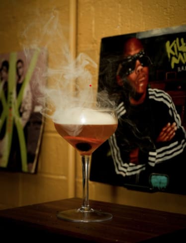 MLB ALL-STAR RESTAURANT WEEK 16 A cocktail with a smoky top layer sits on a wooden table, with a hip-hop album cover in the background in Atlanta, Georgia.