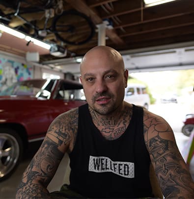 Dr. Dax 1 Tattooed man in tank top sits in garage near vintage red car in Atlanta, Georgia.