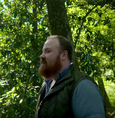 Chef Kevin Gillespie 1 A bearded man in a green vest stands in a lush, green forest in Atlanta, Georgia.