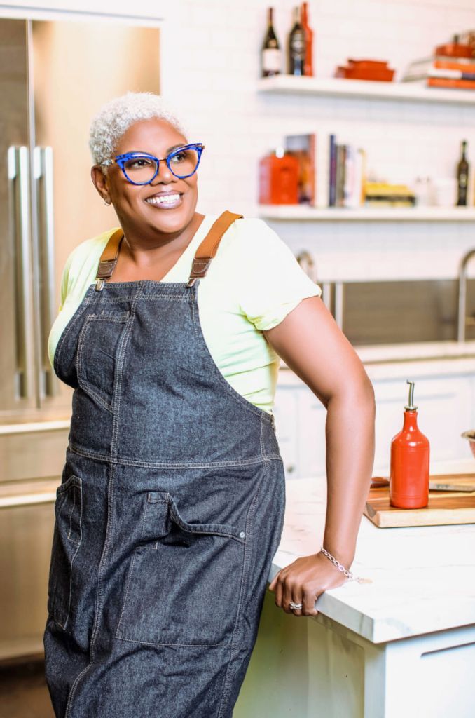 A woman with short white hair, wearing blue-framed glasses, a yellow t-shirt, and denim overalls, stands smiling in a modern kitchen with white cabinets and red accents in Atlanta, Georgia.