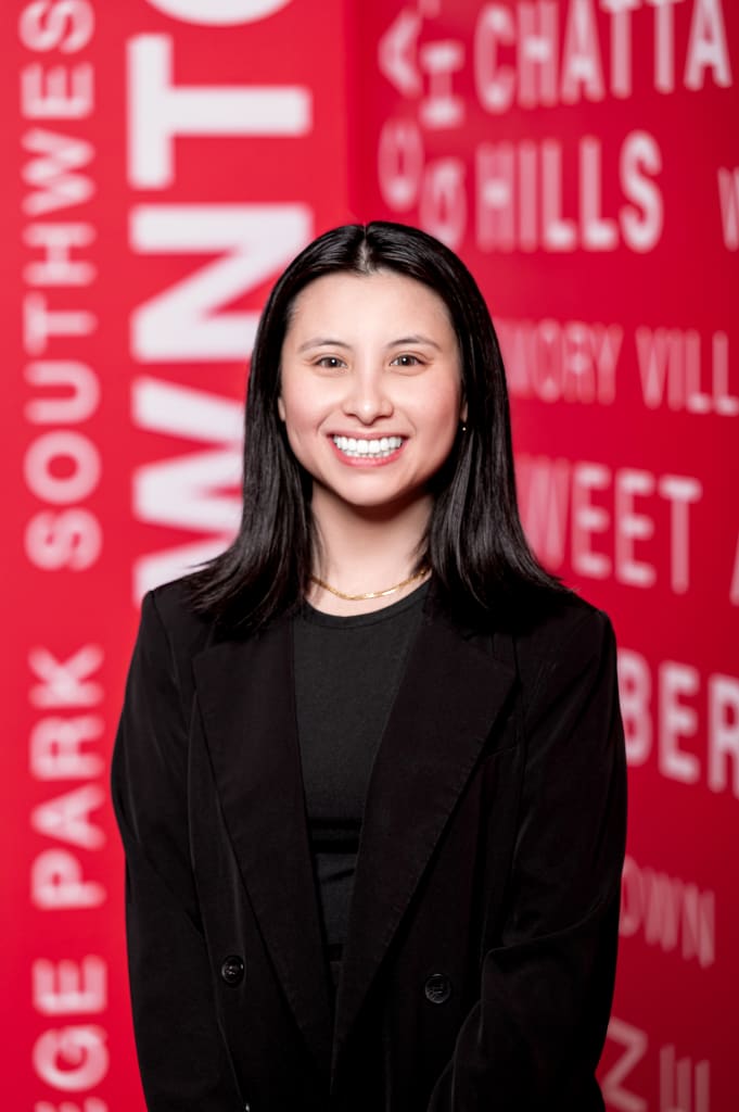 ACVB Promotes LyLy Nguyen to Data and Analytics Manager 1 A smiling woman in a black suit stands in front of a red background with white text in Atlanta, Georgia.