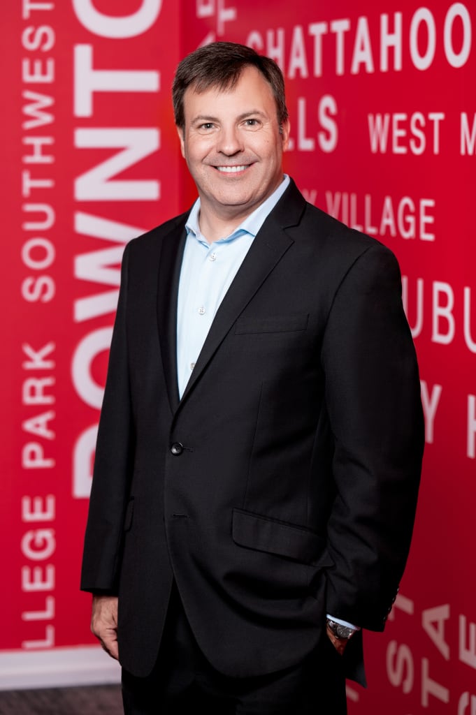 ACVB Promotes Brandon Barnes to Senior Director, International Tourism Sales 1 A man in a black suit stands smiling in front of a red wall with white text in Atlanta, Georgia.