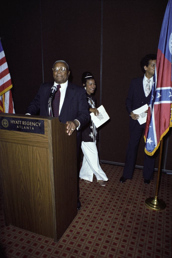 A man stands at a podium labeled 'Hyatt Regency Atlanta' with an American flag and a Confederate flag in the background, while two other individuals hold papers nearby in Atlanta, Georgia.