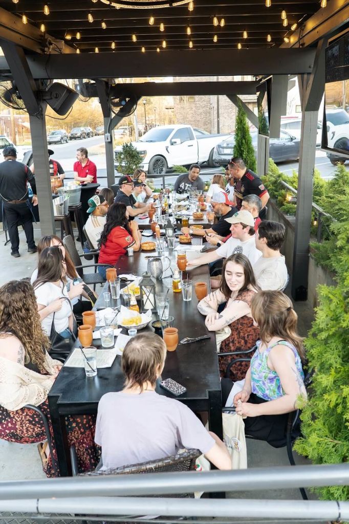 A lively outdoor dining scene with a large group of people gathered around a long table, enjoying a meal together under a string-light canopy in Atlanta, Georgia.