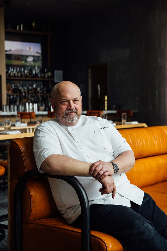 A bald man with a beard, wearing a white chef's jacket, sits on an orange couch in Atlanta, Georgia.