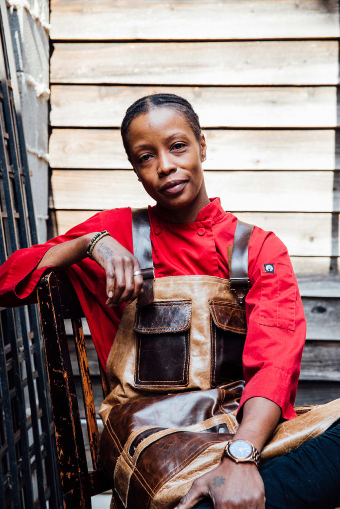 A woman in a red chef's jacket and leather apron sits confidently on a wooden chair in Atlanta, Georgia.
