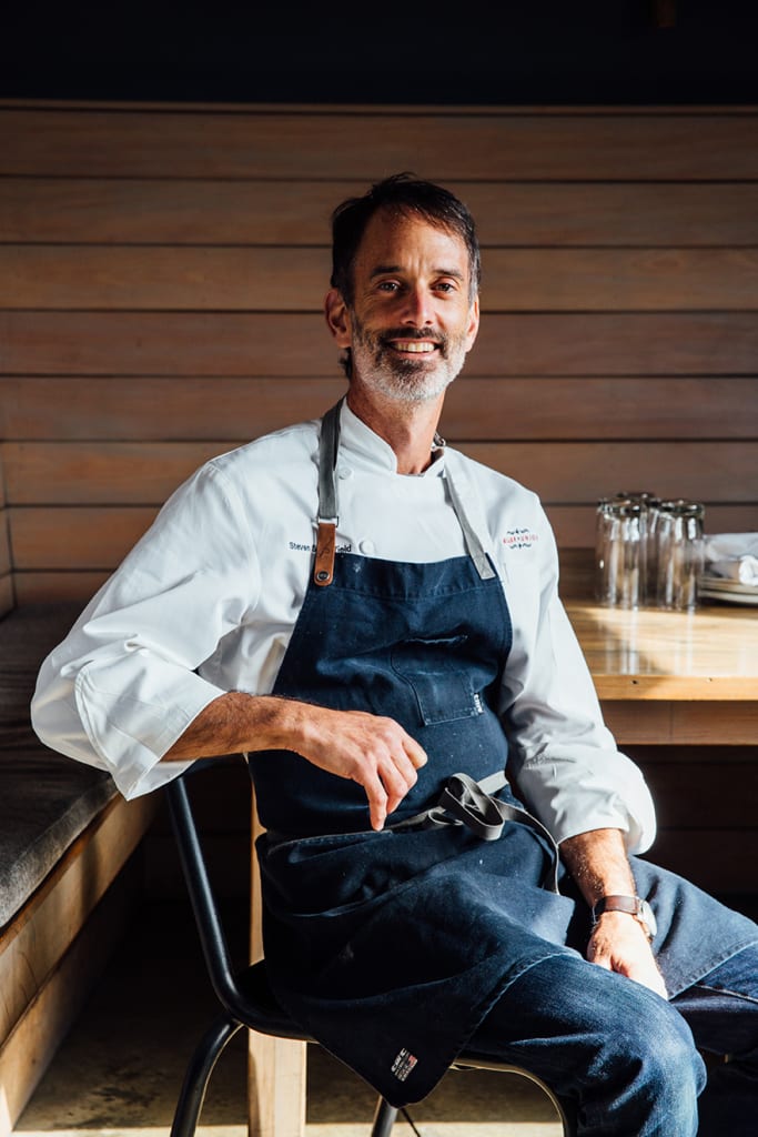 A smiling chef in a white coat and apron sits at a wooden table in Atlanta, Georgia.