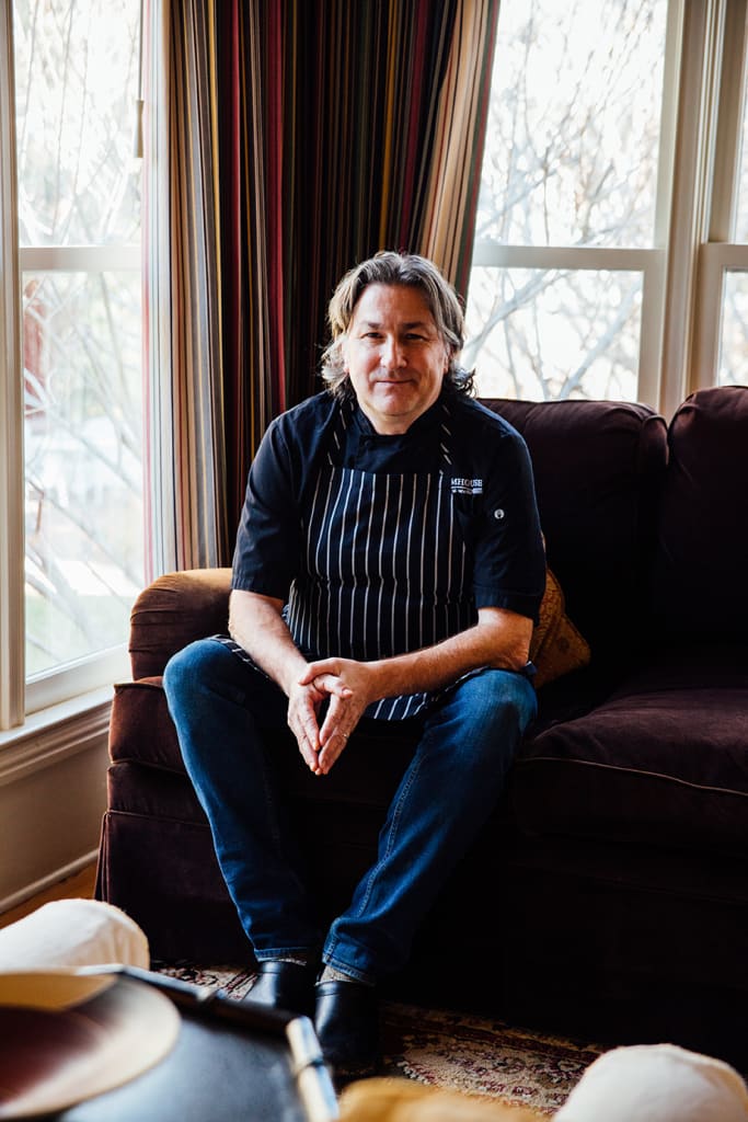 A man in a chef's apron sits on a brown couch near a window with striped curtains in Atlanta, Georgia.