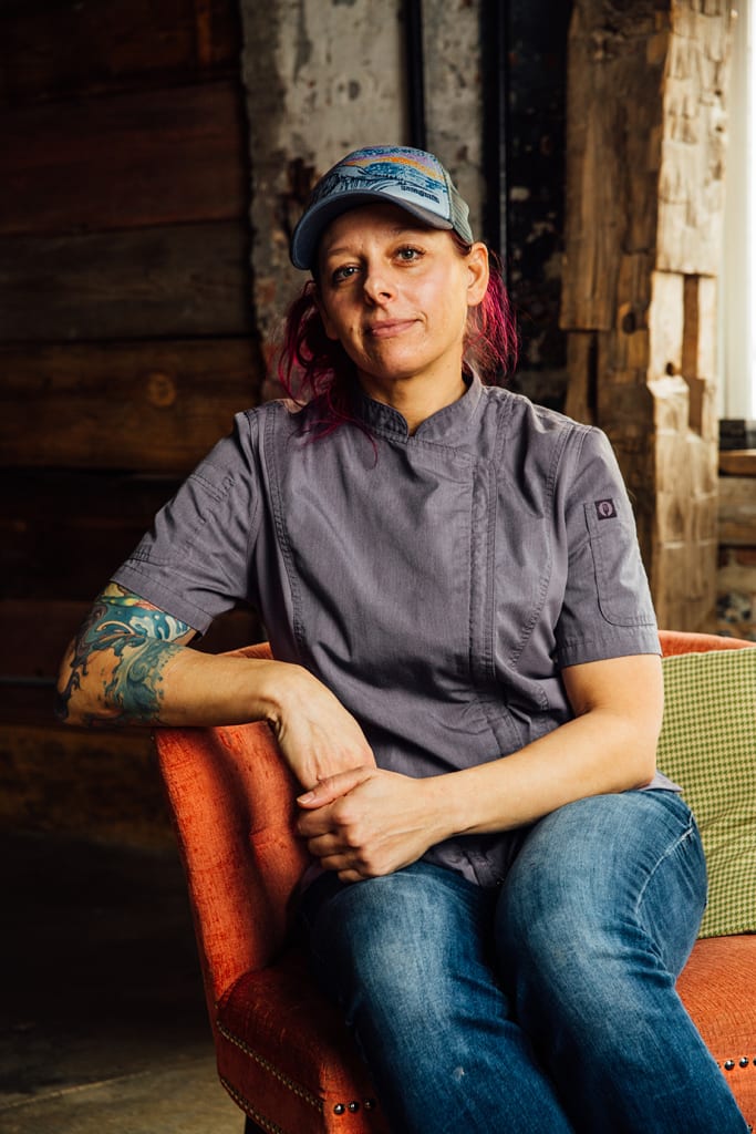 A woman with red hair and a tattooed arm sits on an orange couch wearing a chef's jacket and a hat in Atlanta, Georgia.