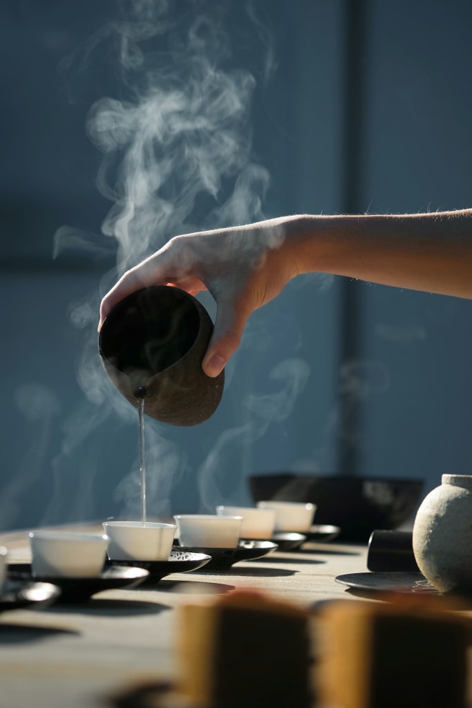 A hand pours steaming tea from a black teapot into a row of white cups on a wooden table in Atlanta, Georgia.