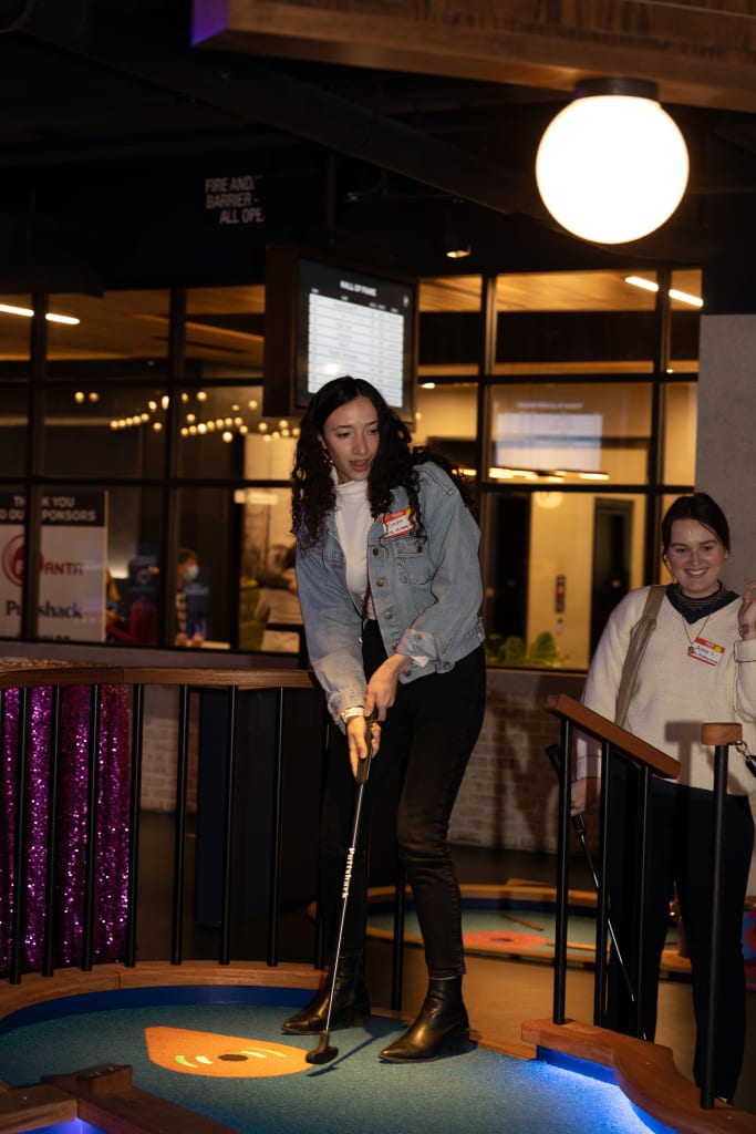 Two women play mini-golf indoors, one wearing a denim jacket and the other a white sweater in Atlanta, Georgia.