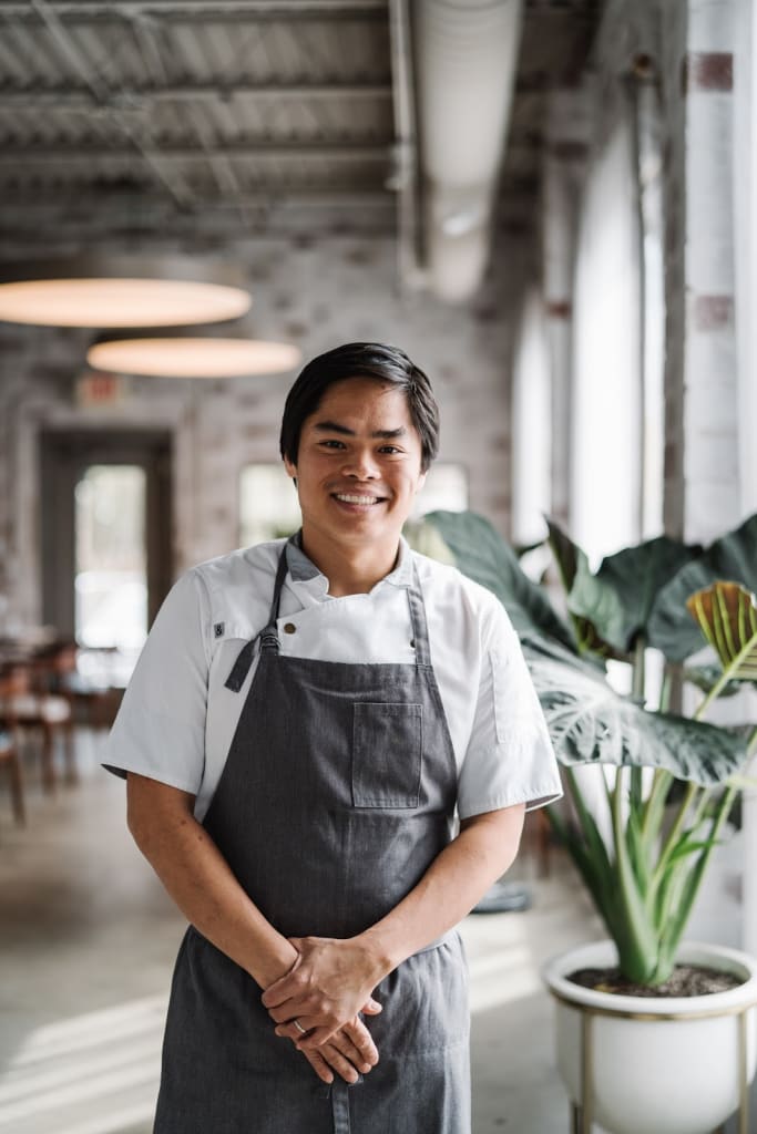 A smiling chef in a white shirt and gray apron stands in a bright, plant-filled kitchen in Atlanta, Georgia.