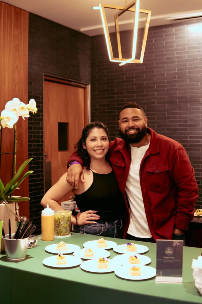 La historia detrás de Bar ANA en Eastside, una carta de amor a la cultura, la artesanía y la comunidad 5 A smiling couple stands behind a table with small plates of food and a flower arrangement in Atlanta, Georgia.
