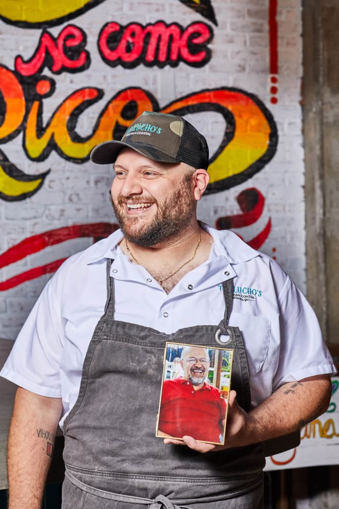 A smiling chef in a white shirt and apron holds a framed photo of a smiling man in a red shirt in Atlanta, Georgia.