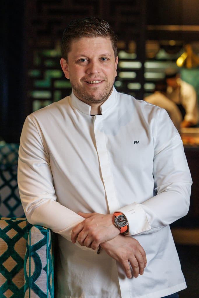 A man in a white chef's jacket smiles confidently while leaning on a patterned armchair in Atlanta, Georgia.