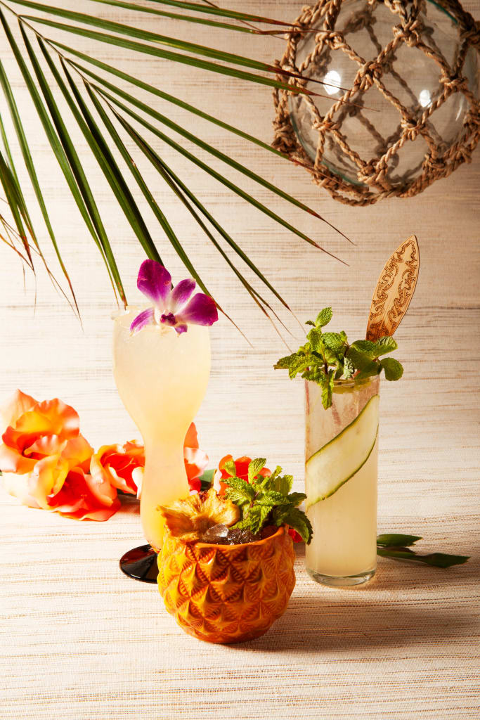 Three tropical cocktails garnished with flowers and herbs, served in a pineapple cup, a tall glass, and a glass with a wooden stirrer, set against a backdrop of palm leaves and a woven net in Atlanta, Georgia.