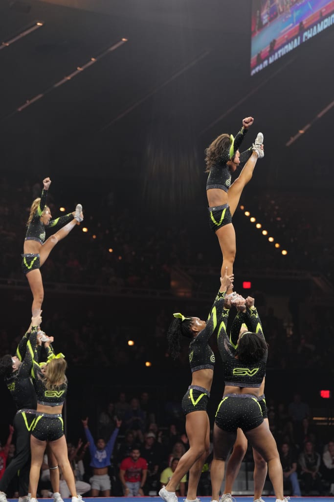 A group of cheerleaders perform a daring human pyramid, with one cheerleader being lifted high into the air by her teammates, all wearing matching black and neon green uniforms in Atlanta, Georgia.